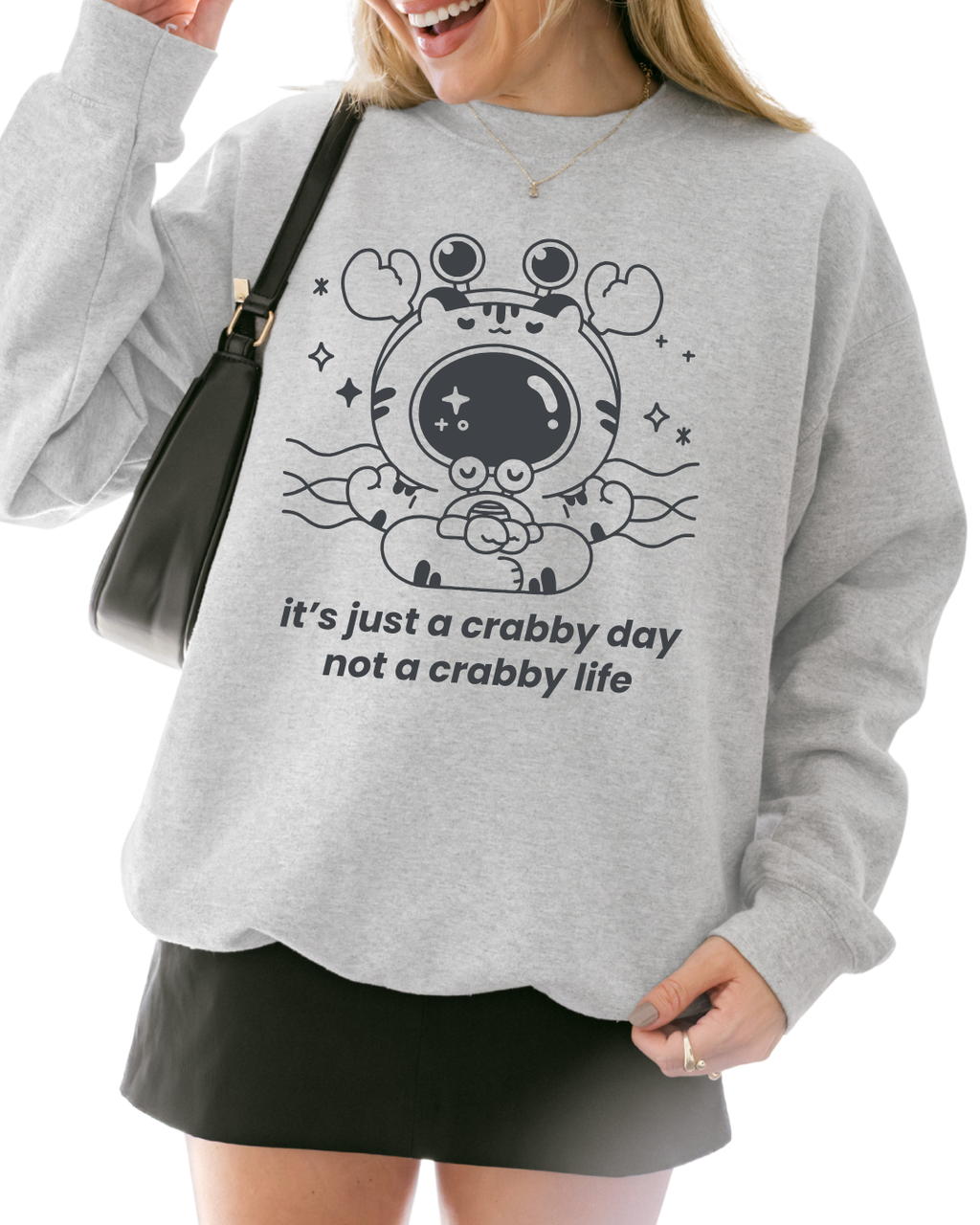 Person wearing an ash gray graphic crewneck sweatshirt with a meditating crab illustration and the phrase “it’s just a crabby day, not a crabby life”