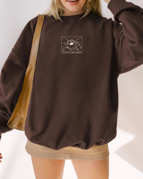 Model wearing a chocolate brown embroidered crewneck sweatshirt with a small tiger illustration and the phrase “It’s OK to Take a Break,” styled casually for everyday wear.