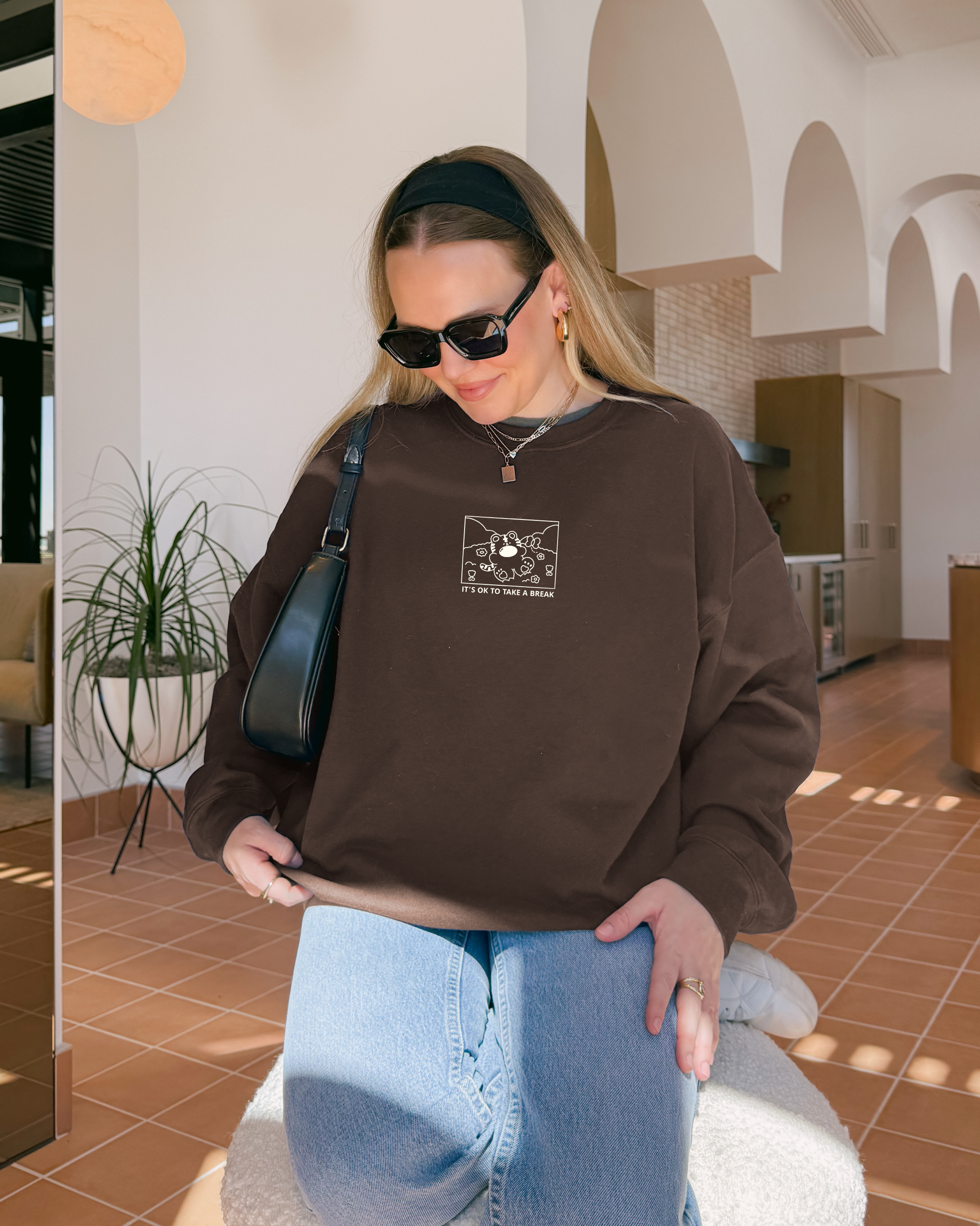 Model wearing a chocolate brown embroidered sweatshirt with the phrase “It’s OK to Take a Break,” styled indoors for a calm, rest-focused lifestyle.