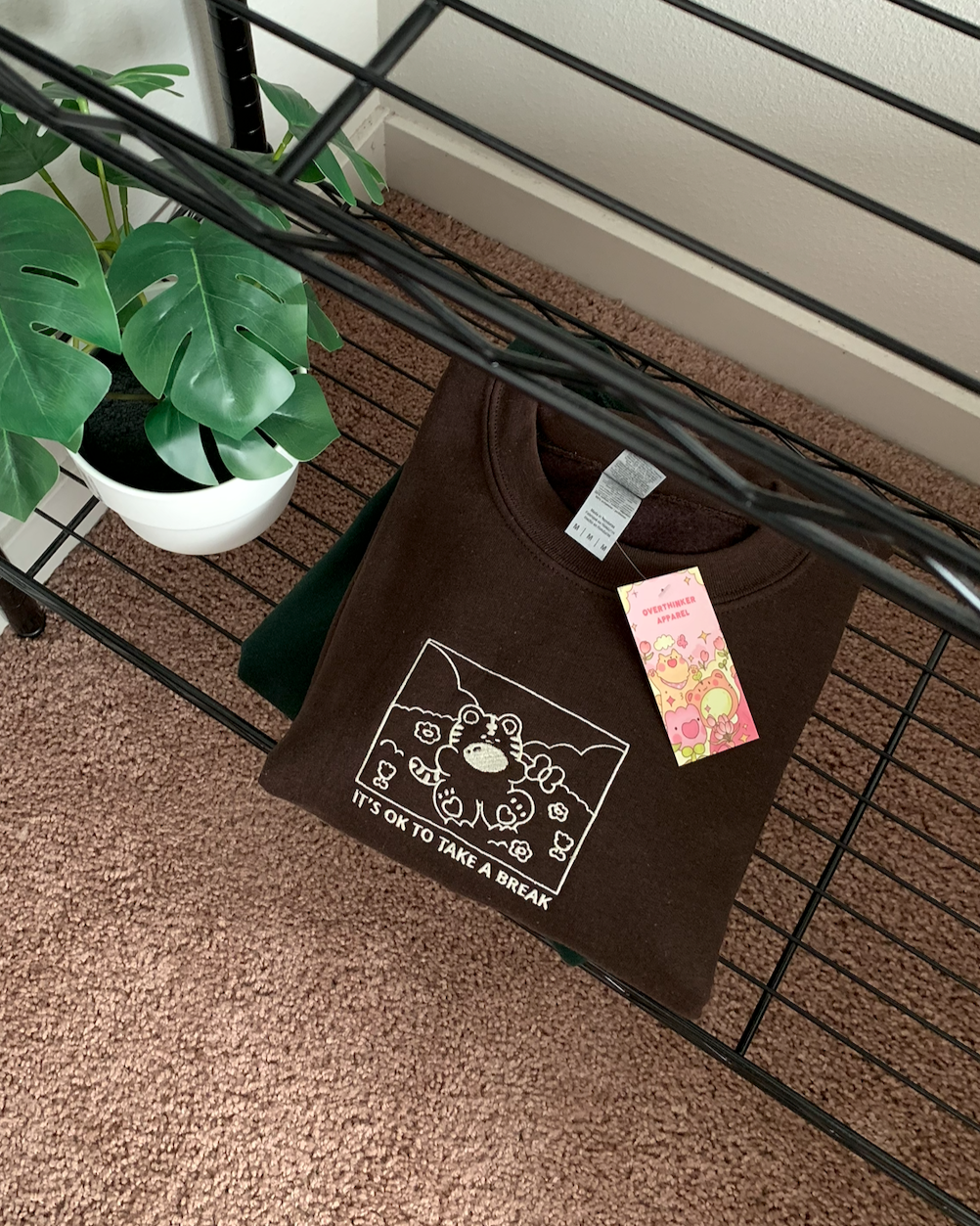 Brown ‘It’s OK to take a break’ sweatshirt folded neatly on a black shelf with plant and Overthinker Apparel hang tag visible