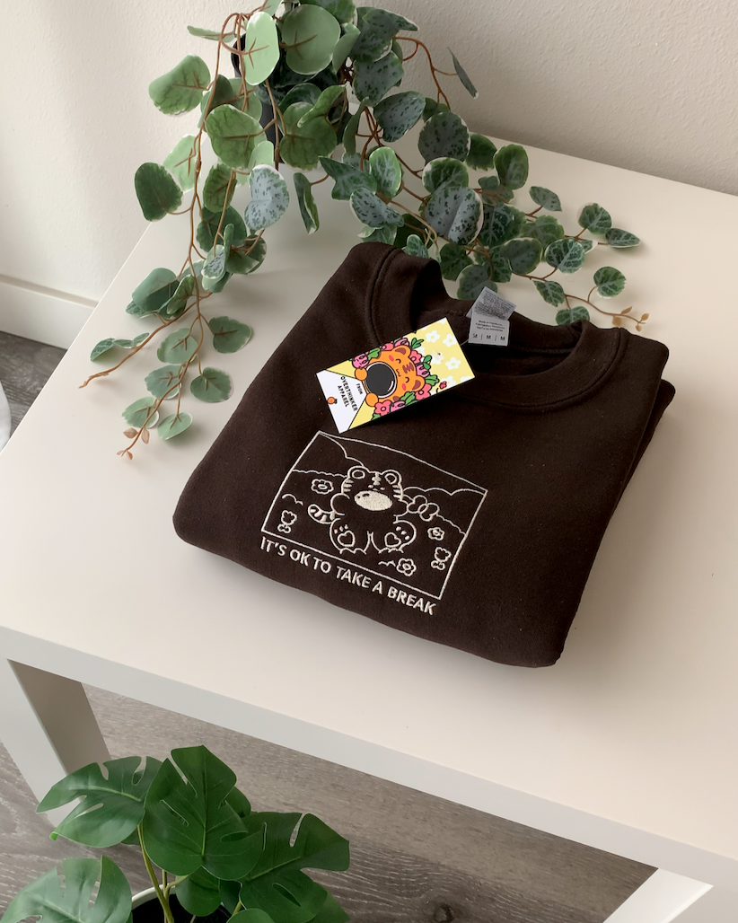 Folded brown sweatshirt with ‘It’s OK to take a break’ embroidery and Overthinker Apparel tag, styled with plants on a white table