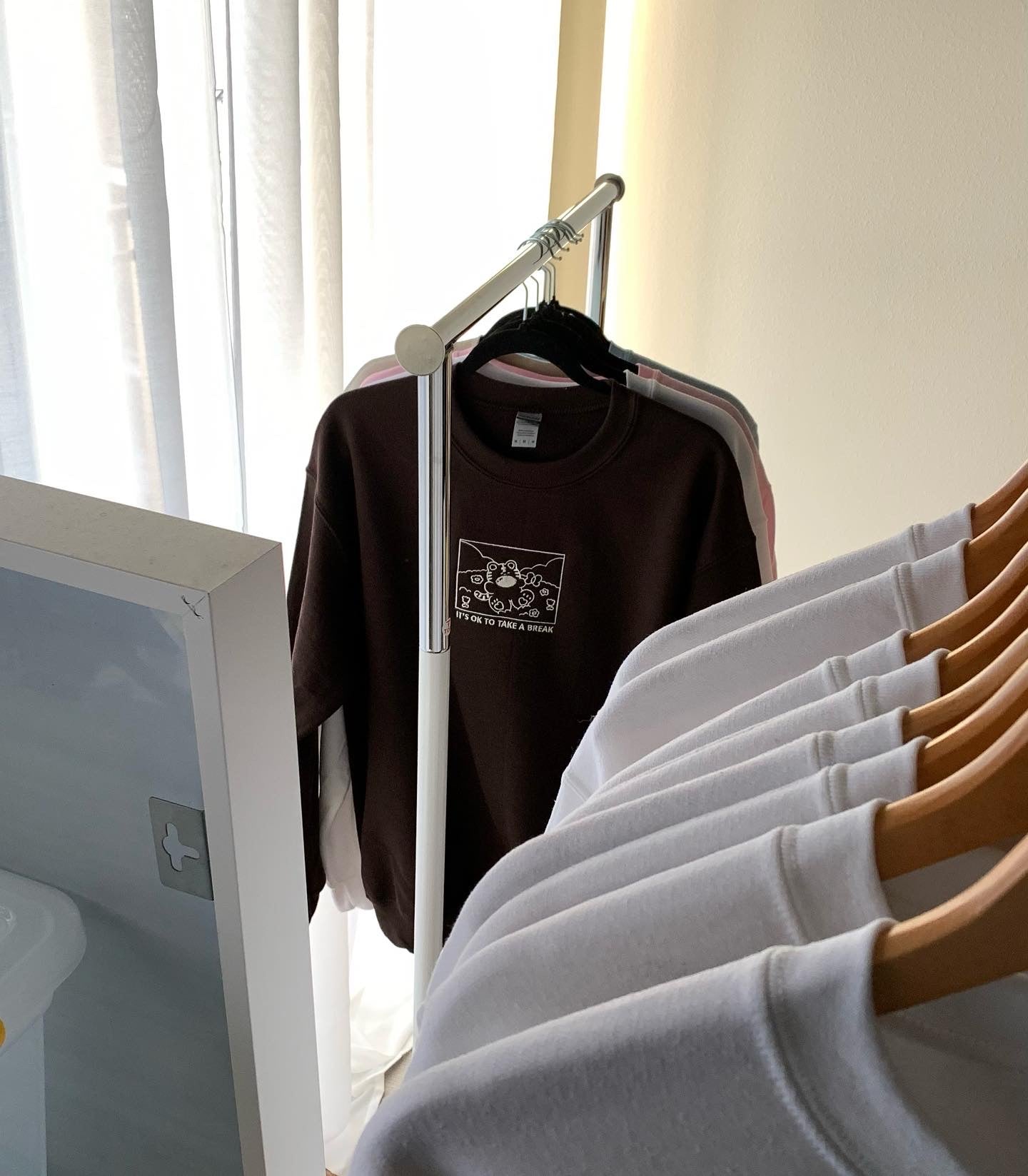 Brown crewneck sweatshirt with ‘It’s OK to take a break’ design hanging on a clothing rack with pastel pieces in natural light