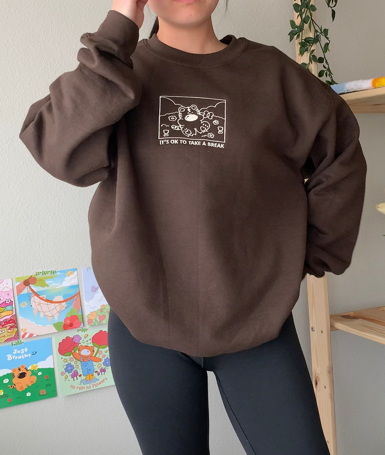 Brown ‘It’s OK to take a break’ sweatshirt modeled indoors with black leggings and colorful art prints on the wall