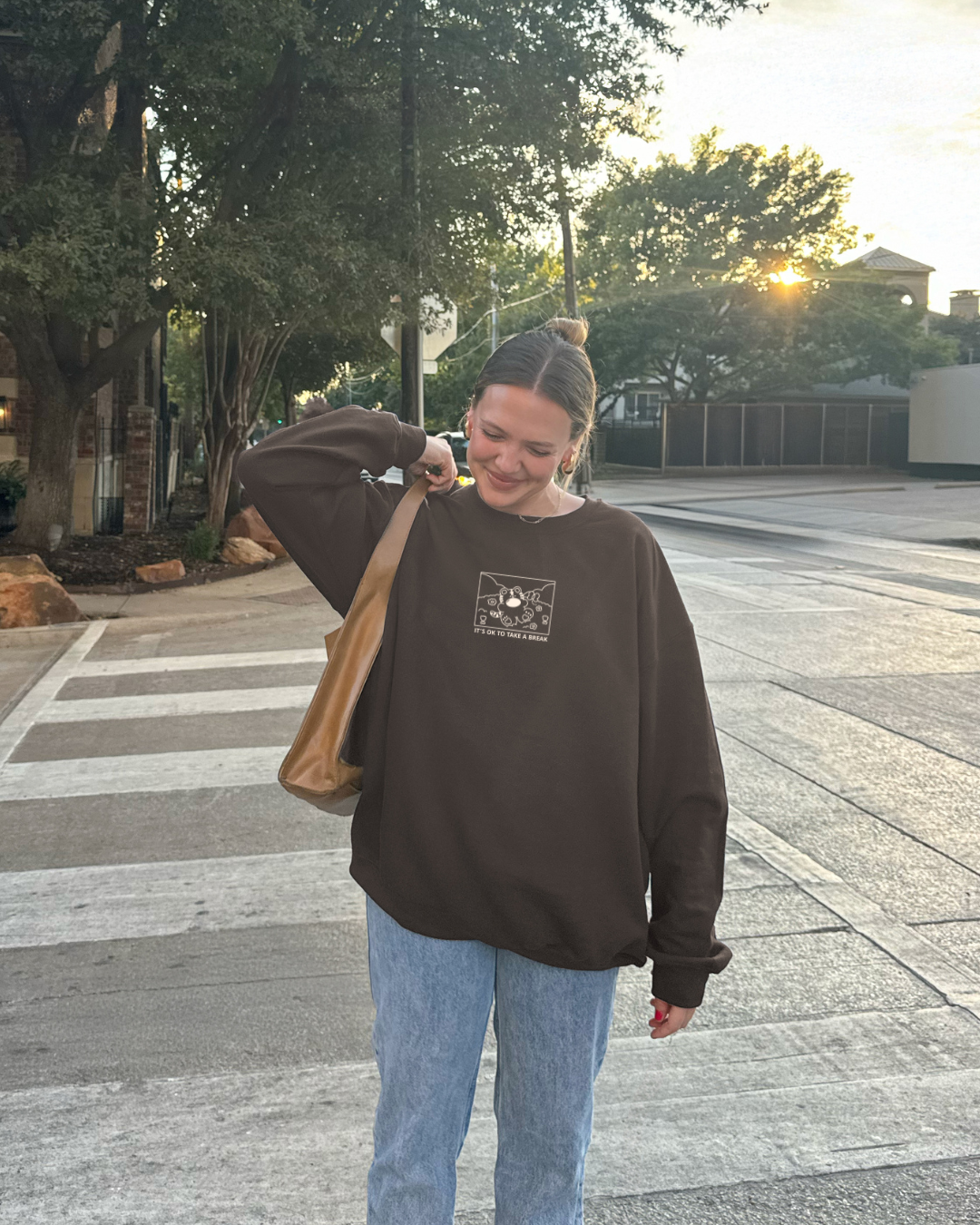 Brown sweatshirt with ‘It’s OK to take a break’ tiger design, worn outdoors with jeans and shoulder bag during golden hour