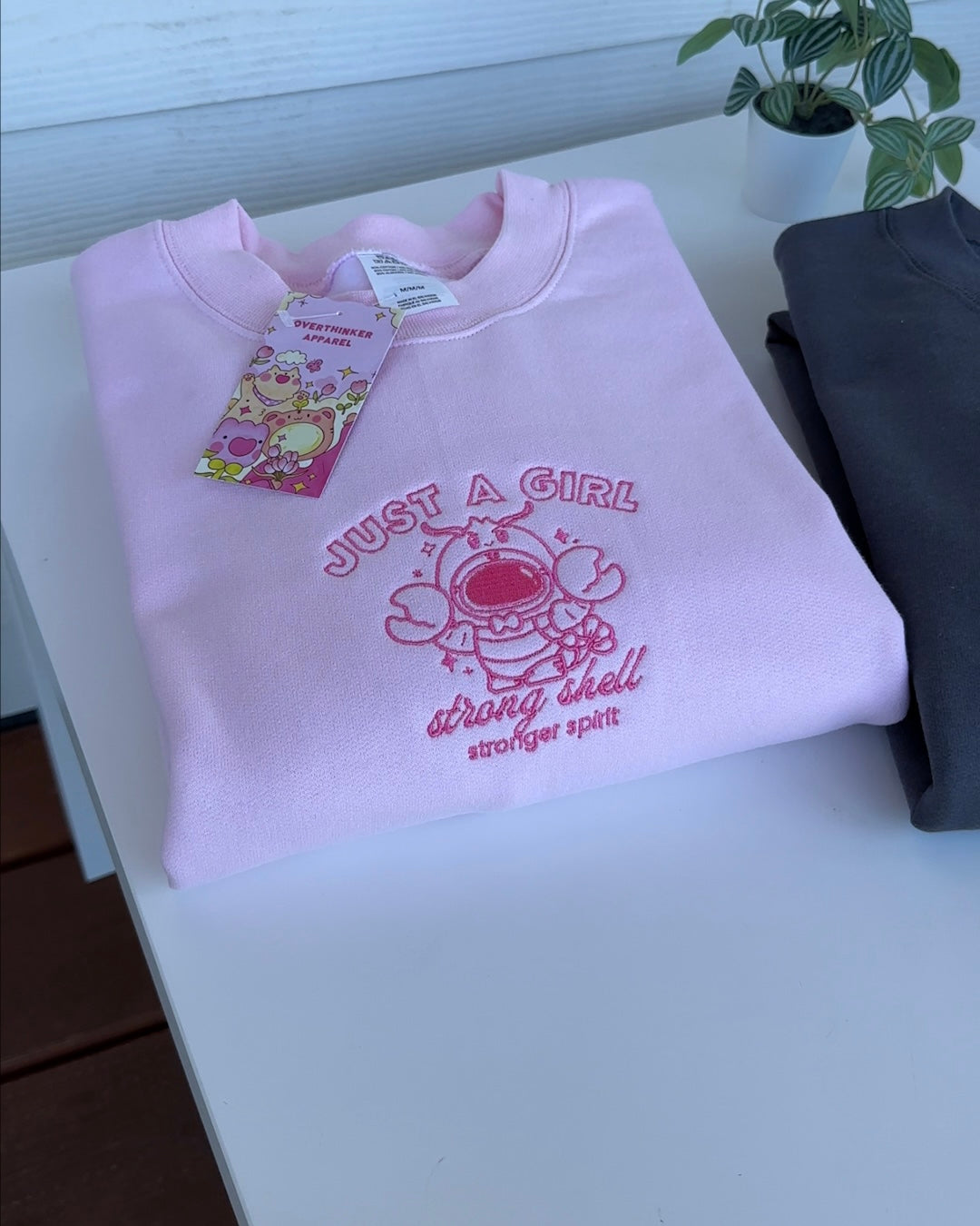 Folded pink embroidered sweatshirt featuring a playful lobster astronaut illustration and “Just a Girl” text.