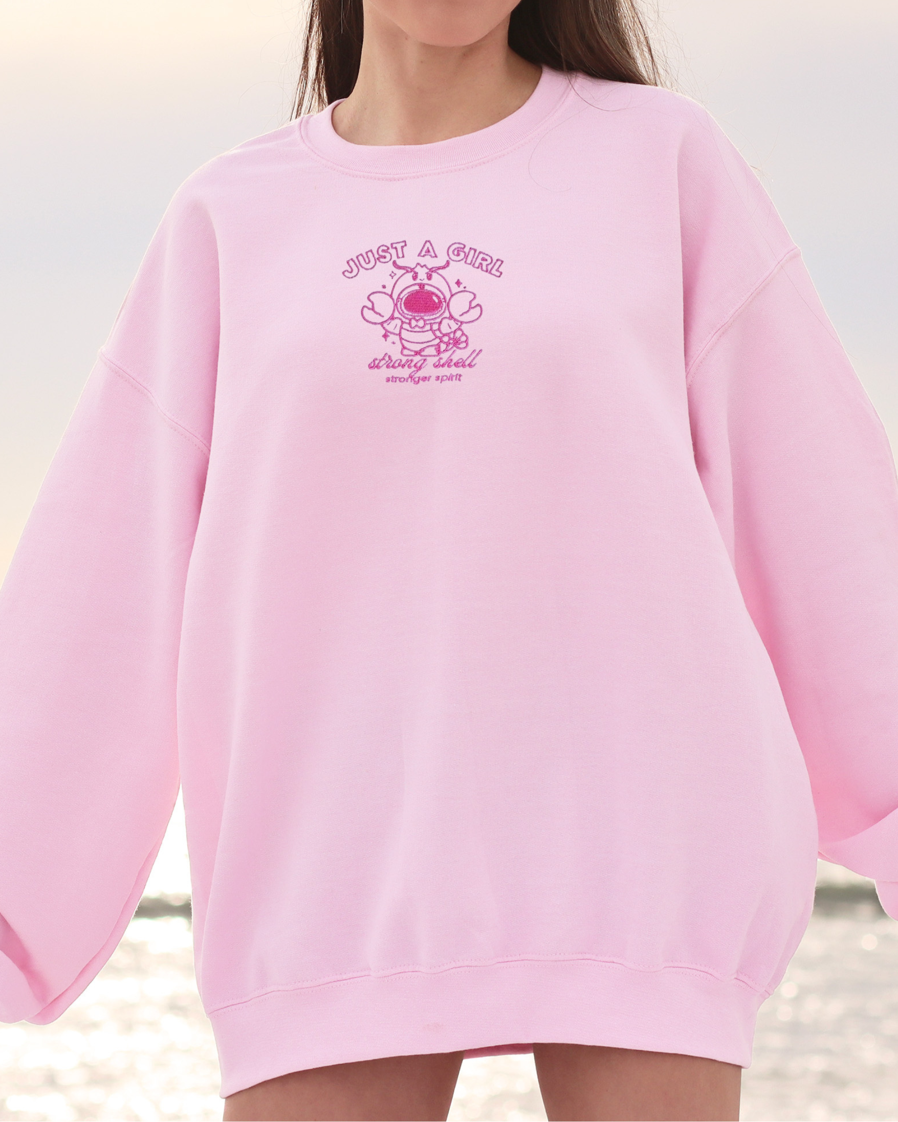 Model wearing a pink oversized embroidered sweatshirt featuring a small lobster astronaut design and the phrase “Just a Girl — strong shell, stronger spirit.”