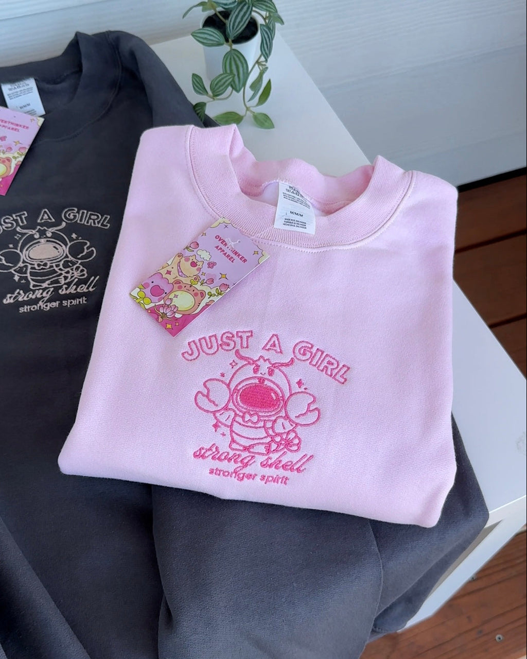 Pink and charcoal embroidered crewneck sweatshirts folded on a table, showing the “Just a Girl” lobster astronaut design.