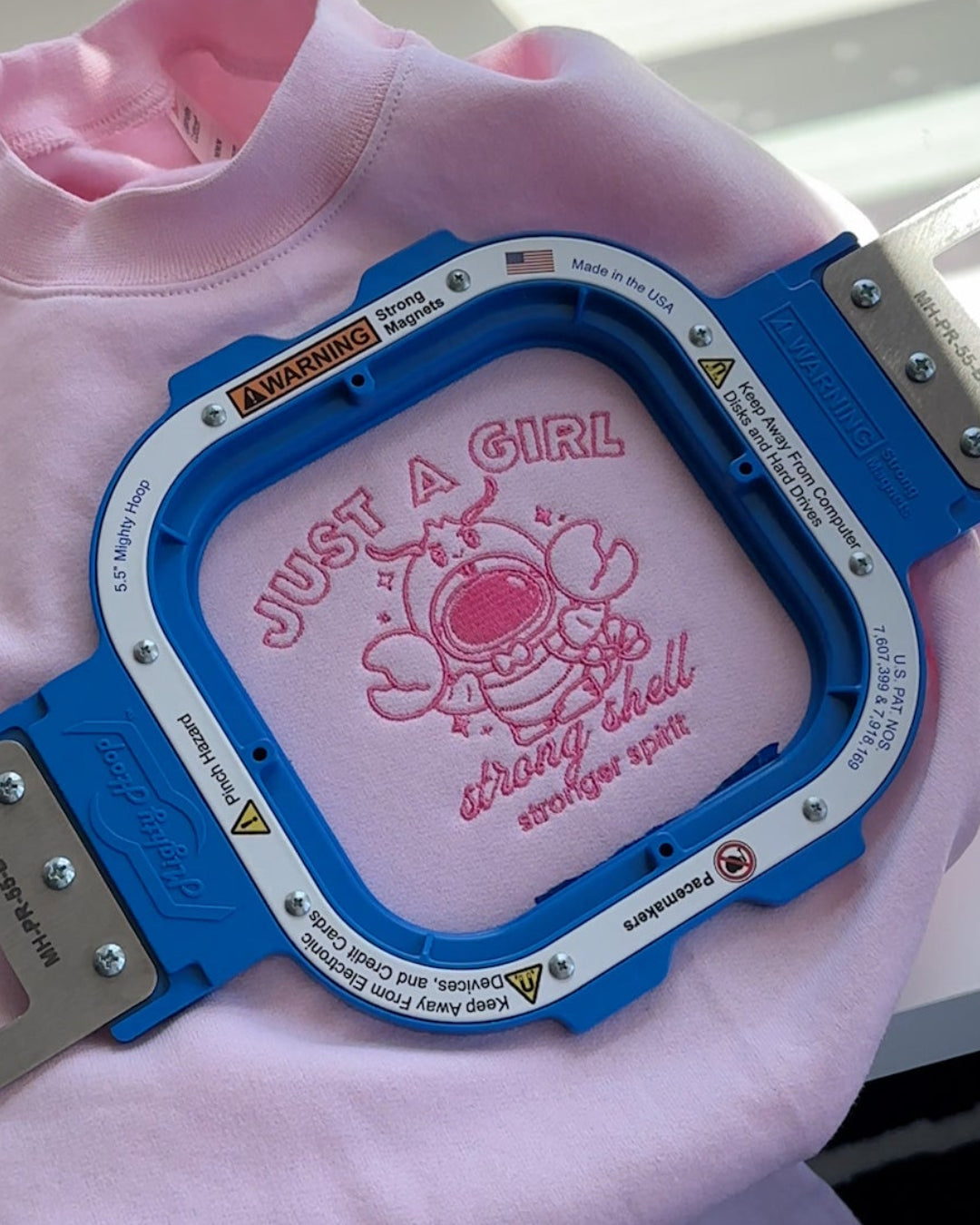 Close-up embroidery of a lobster astronaut with the text “Just a Girl — strong shell, stronger spirit” stitched on a pink sweatshirt.