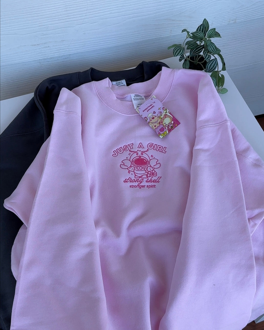 Pink oversized crewneck sweatshirt laid flat with embroidered lobster astronaut artwork and confidence message on the chest.