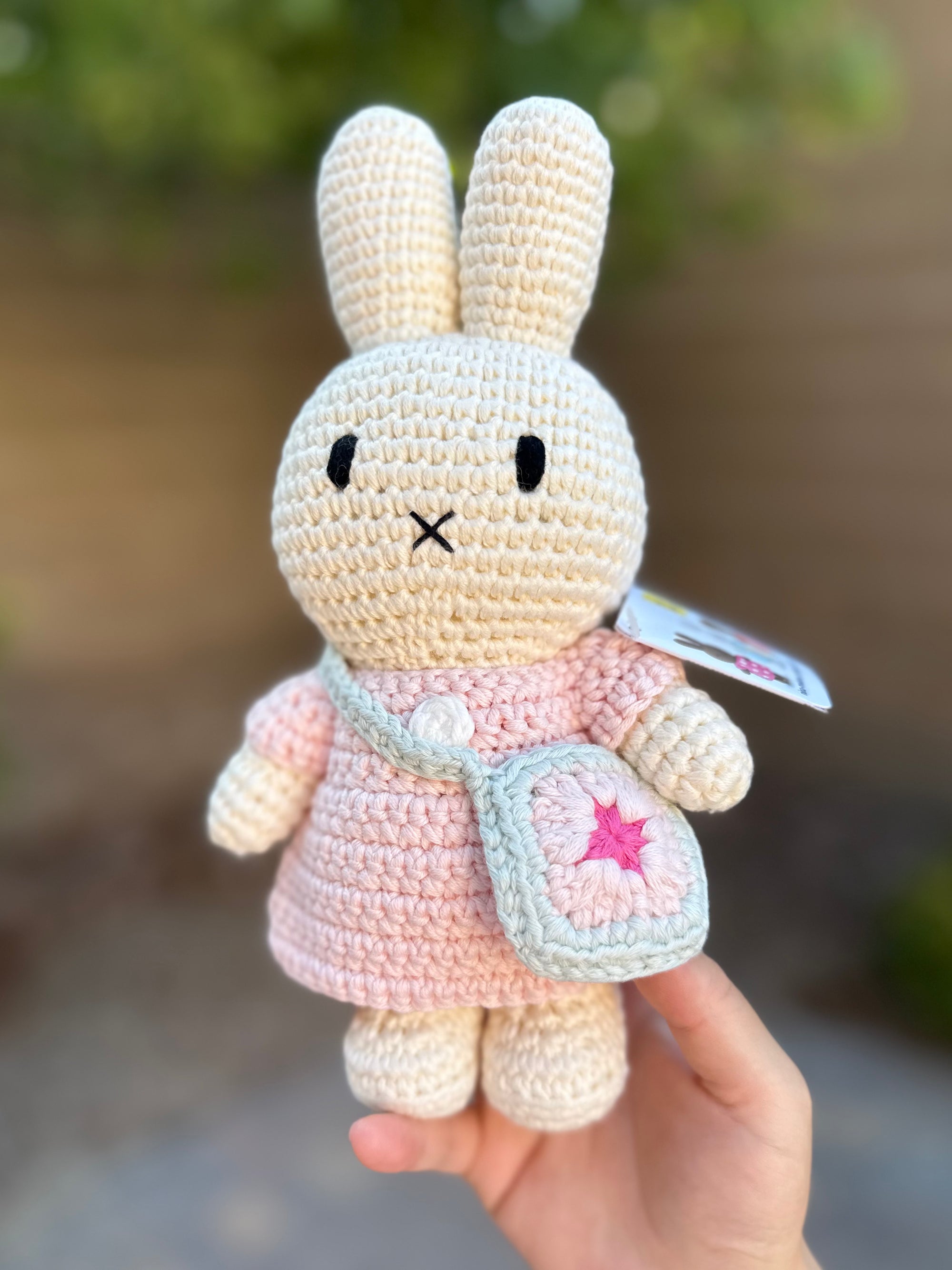 Handmade Miffy crochet plush in a pink dress with removable flower bag, photographed outdoors against greenery.