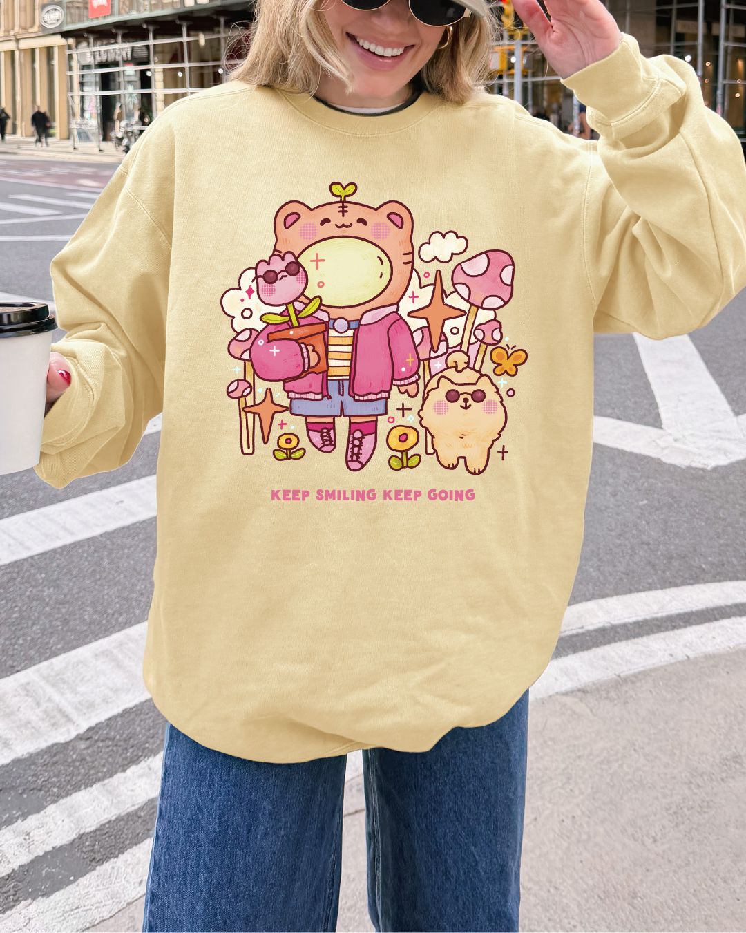 Woman wearing a butter yellow “keep smiling keep going” graphic sweatshirt while holding coffee on a city street, styled as a relaxed everyday outfit