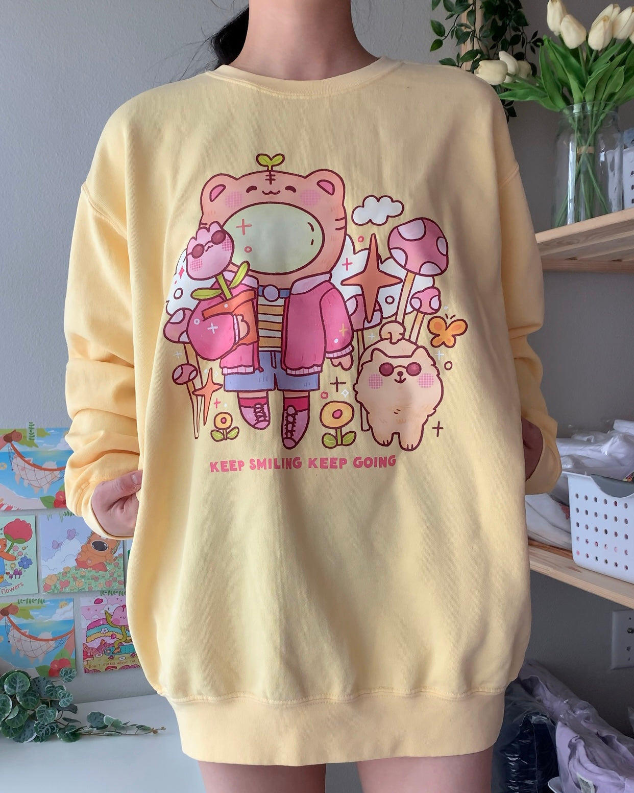 Model wearing a butter yellow graphic crewneck sweatshirt featuring a soft illustrated character design and the phrase “Keep smiling, keep going,” styled for a warm, comforting everyday look.