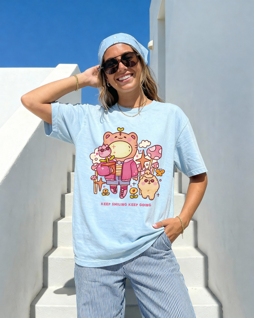 Woman wearing a light blue “keep smiling keep going” graphic tee with a playful illustrated bear design outdoors in bright sunlight