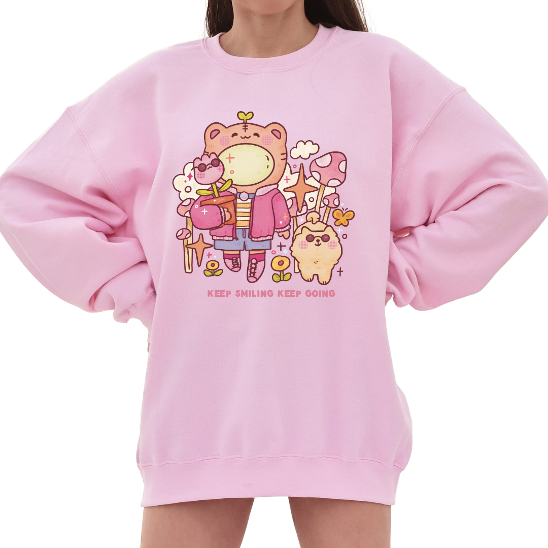 Model wearing a pastel pink graphic crewneck sweatshirt featuring a soft illustrated character design and the phrase “Keep smiling, keep going,” styled for a gentle, comforting everyday look.