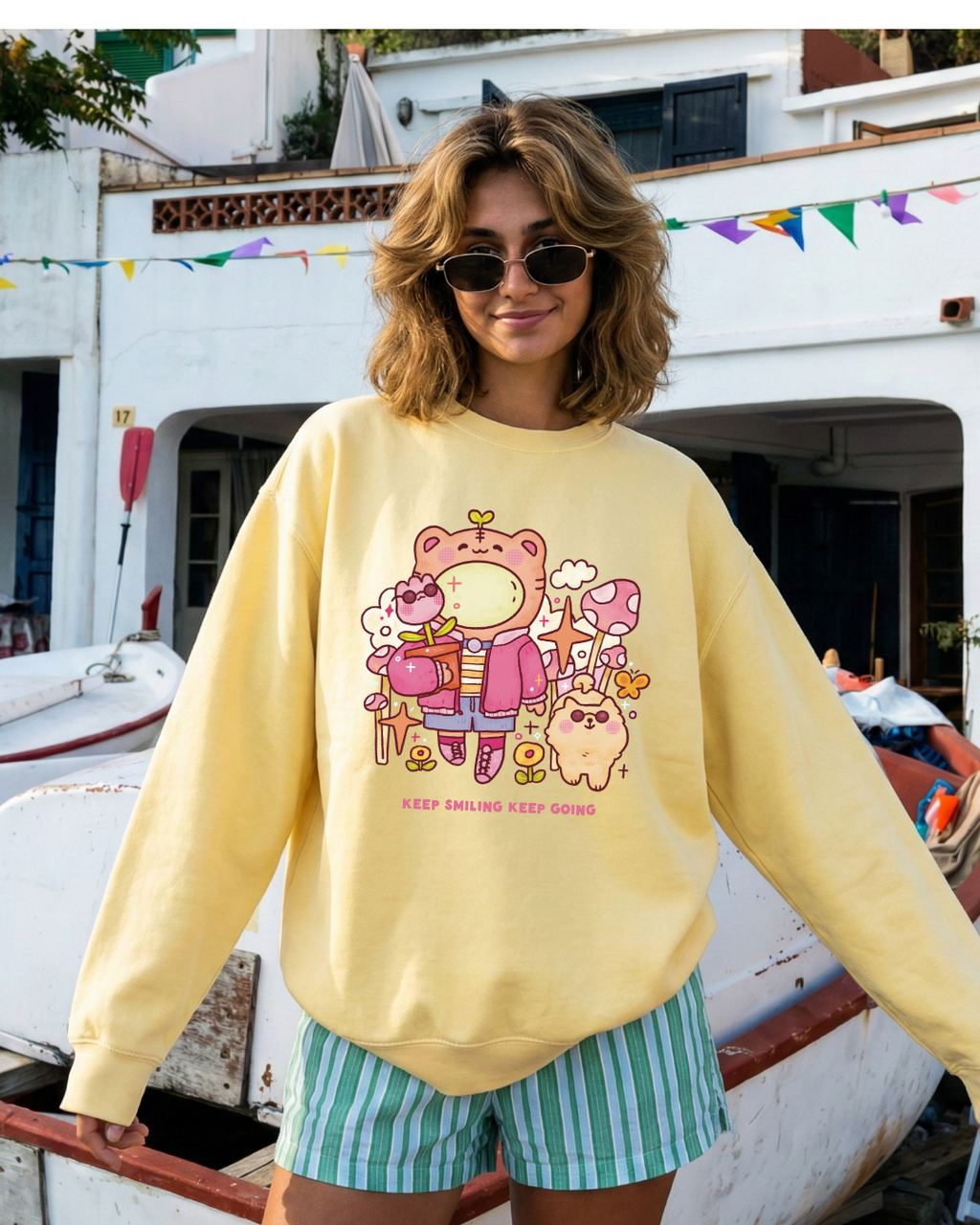 Butter yellow crewneck sweatshirt with a playful illustration and the phrase “keep smiling keep going,” worn casually outdoors in a calm, cozy setting