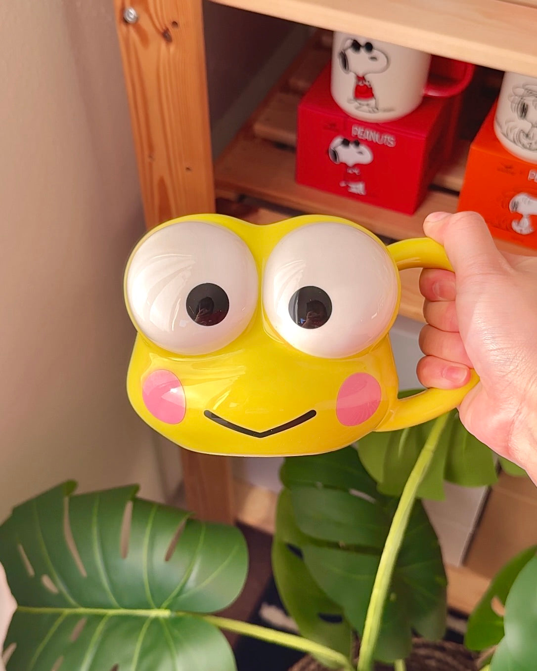 Yellow Keroppi 3D sculpted ceramic mug with wide eyes and pink cheeks, held in hand in a cozy shelf setup with plants and cute decor, officially licensed Sanrio mug.