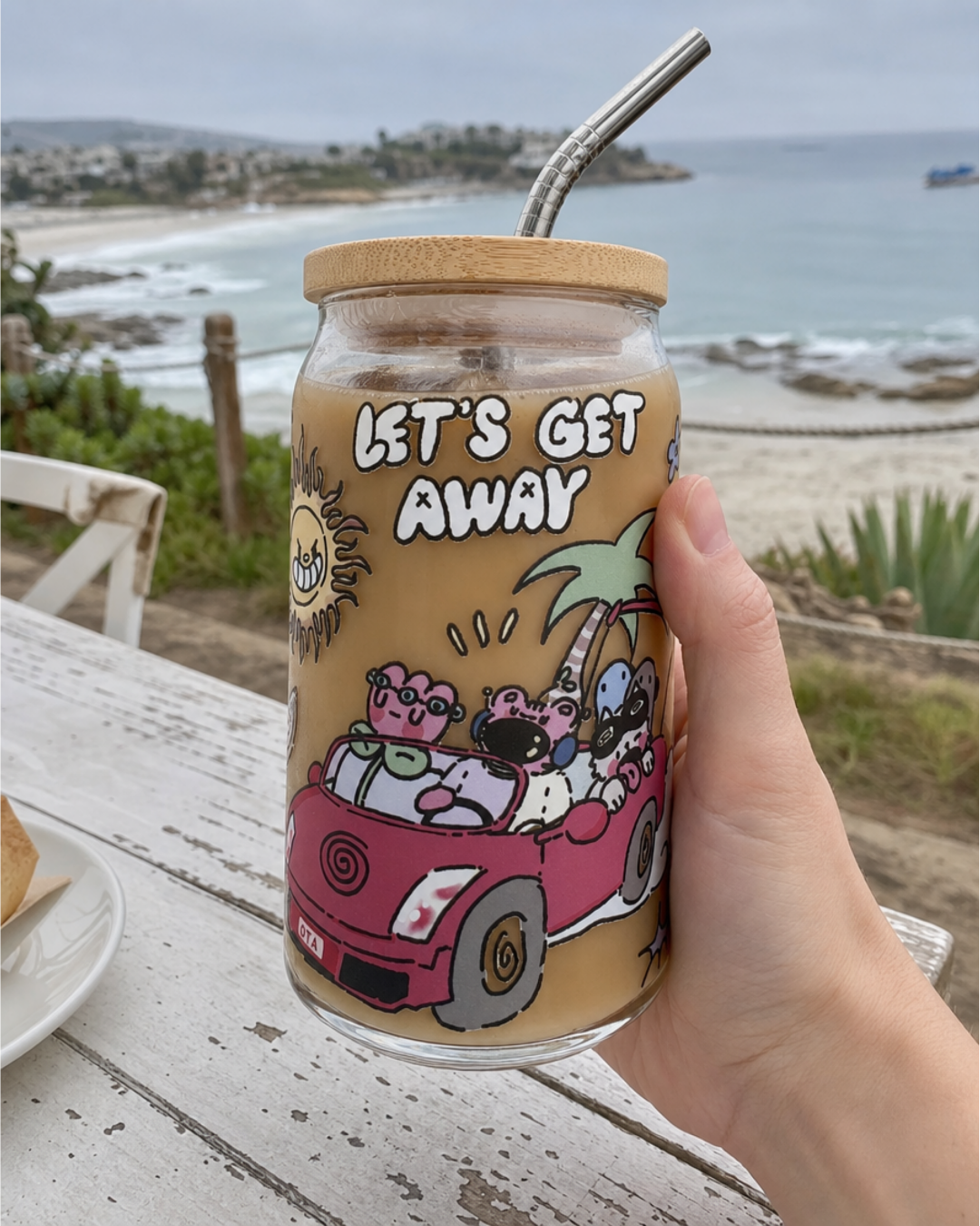 Hand holding the Let's Get Away glass can with a bamboo lid and metal straw, filled with iced coffee, overlooking a beach and ocean view.