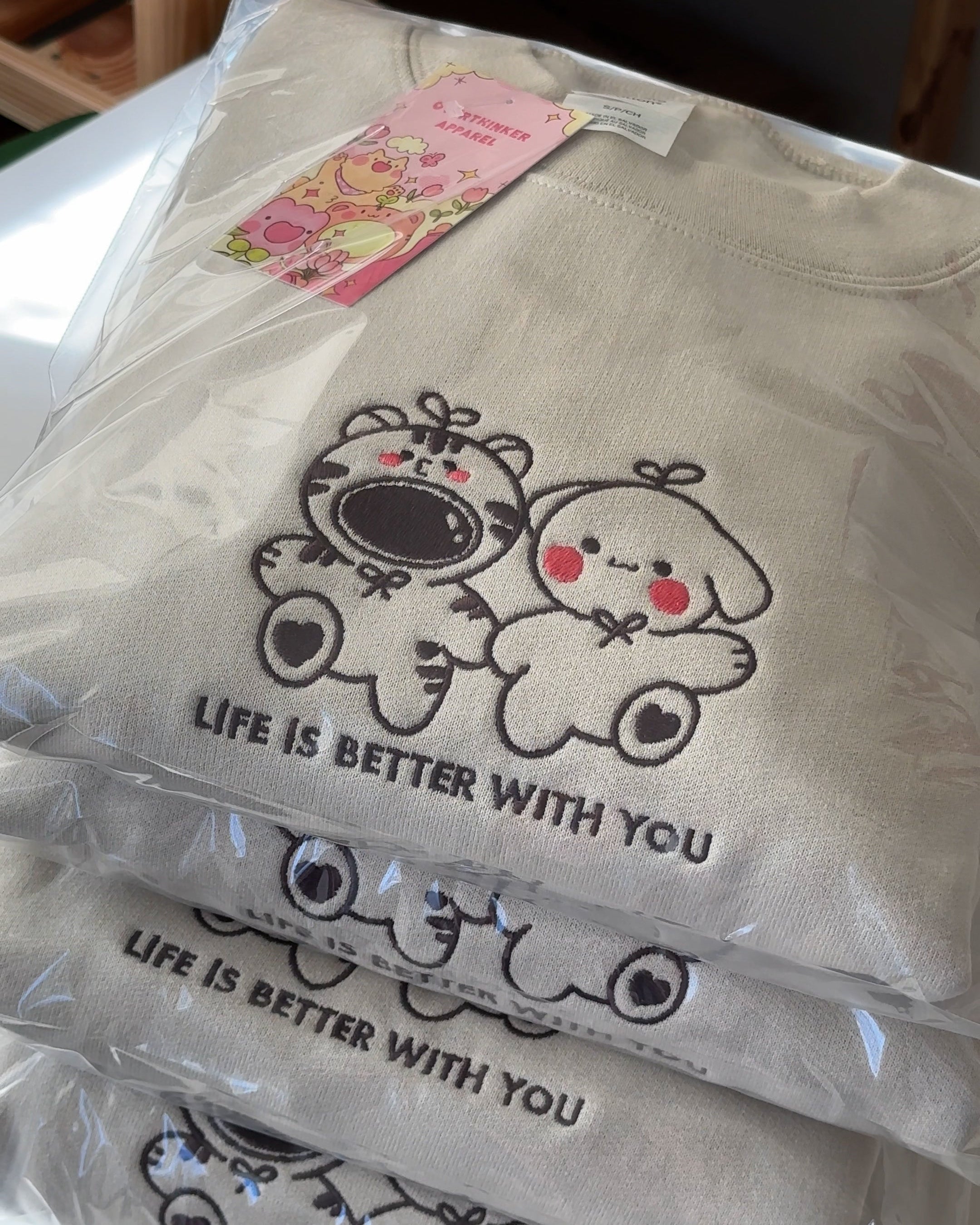 Folded sand beige embroidered sweatshirt featuring two cute characters and the phrase “Life Is Better With You,” shown packaged and ready to ship.