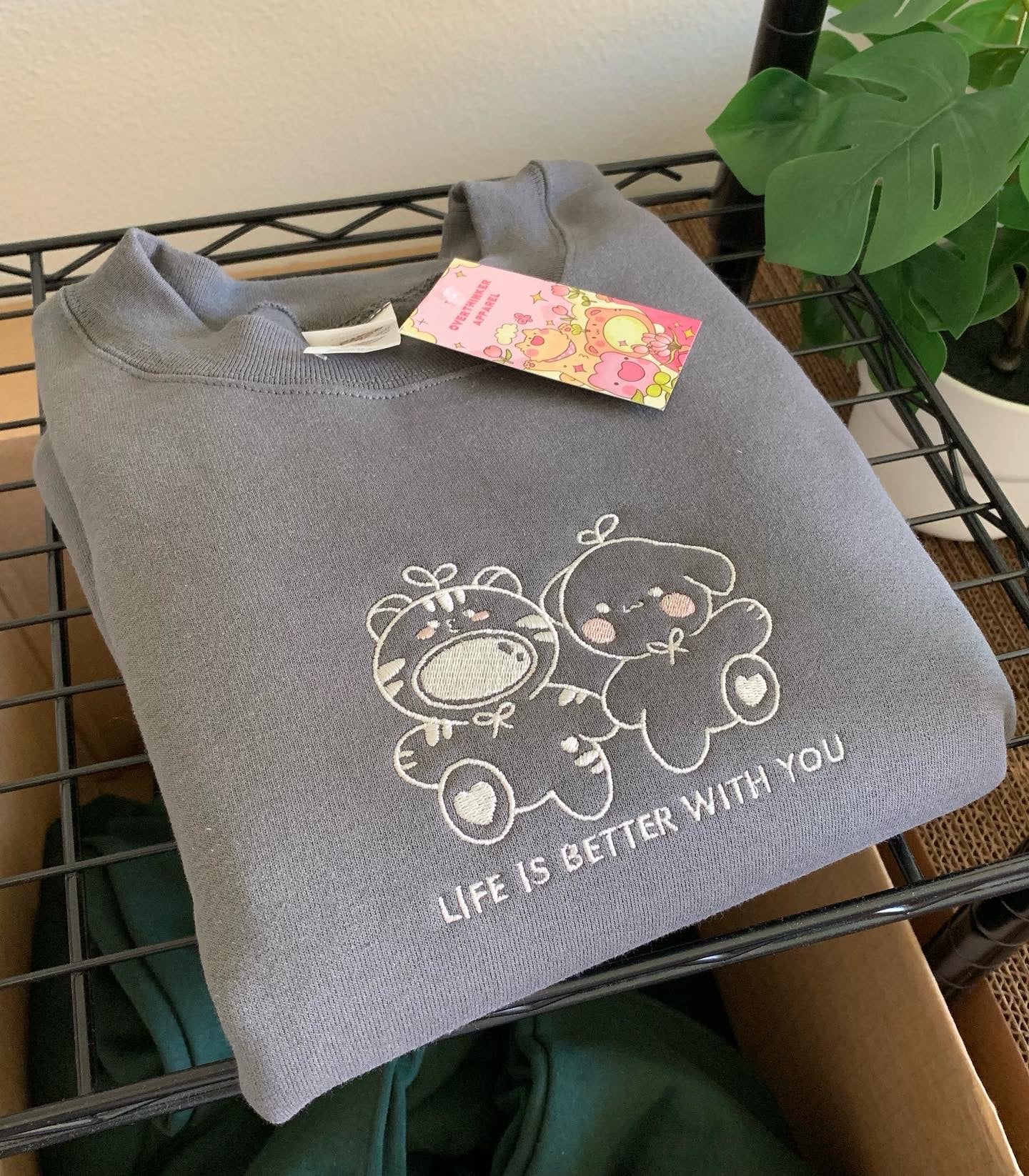Gray sweatshirt with ‘Life is better with you’ embroidered animal duo design, folded on wire shelf with Overthinker Apparel hang tag