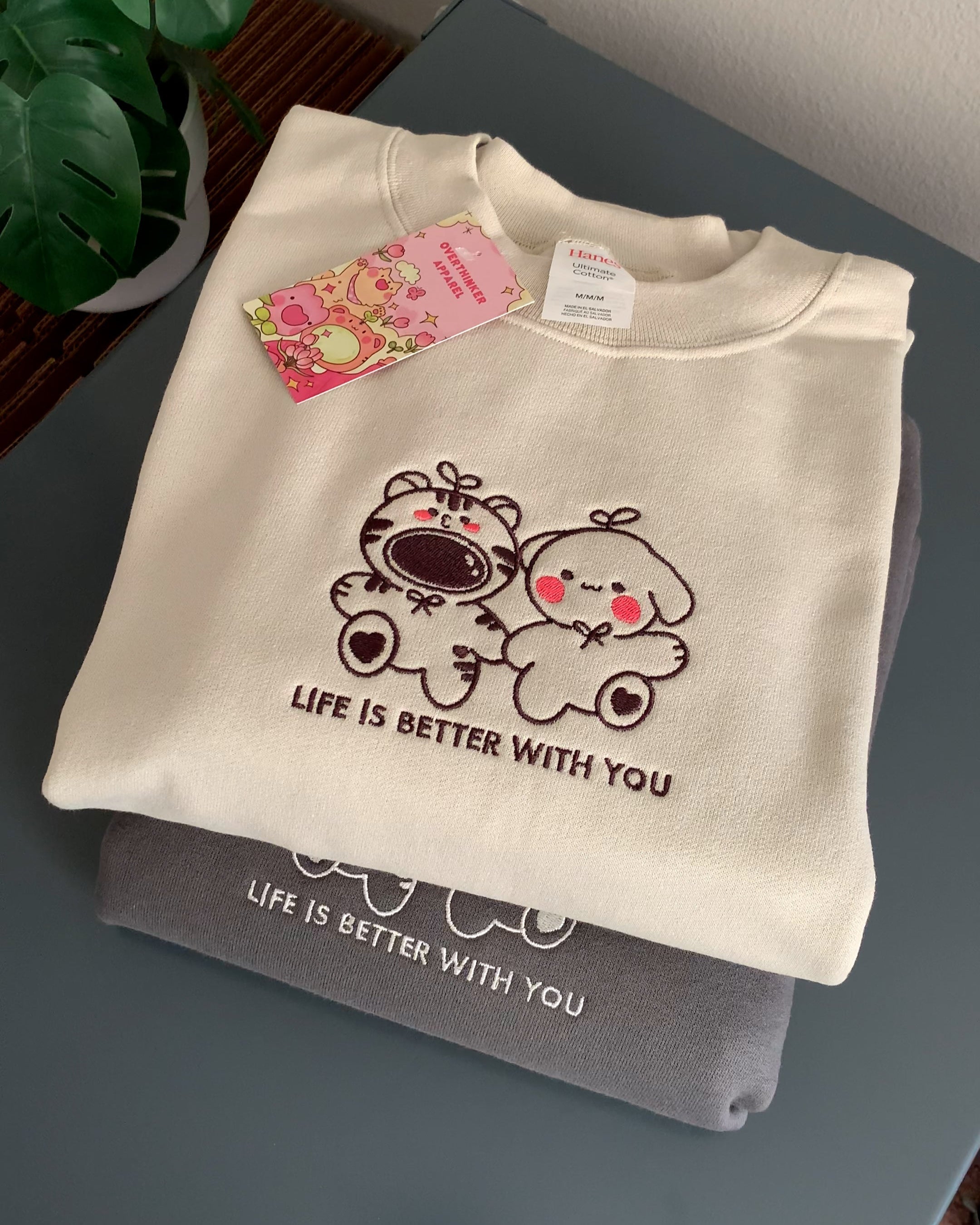 Stack of cream and gray sweatshirts with embroidered ‘Life is better with you’ design, styled with pink hang tag and plant on tabletop