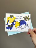 Illustrated greeting card with two people in blue and yellow outfits lounging together, text reads ‘Life is crazy… but I have you,’ paired with light blue envelope.