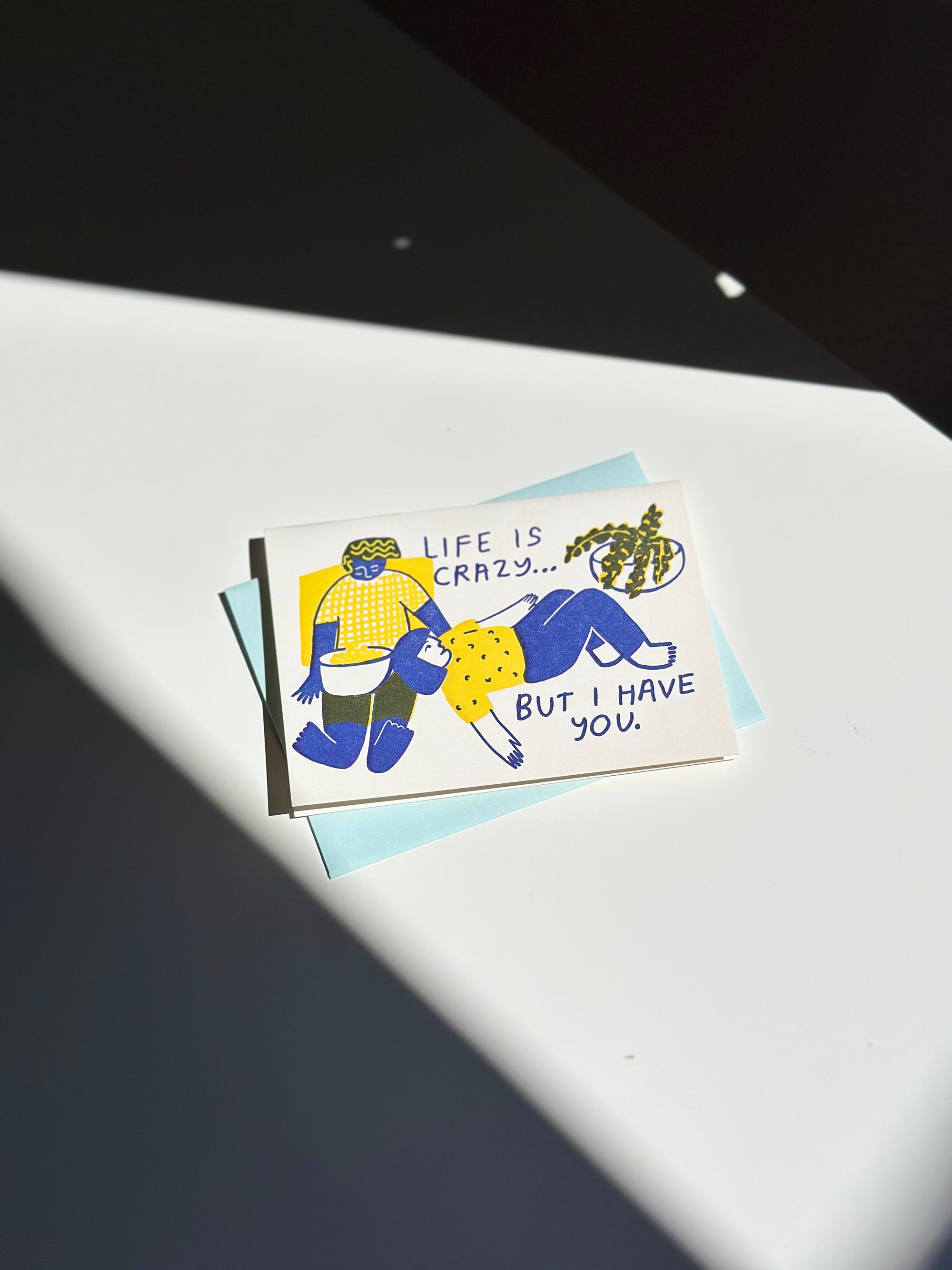 Illustrated greeting card with two people in blue and yellow outfits lounging together, text reads ‘Life is crazy… but I have you,’ paired with light blue envelope.