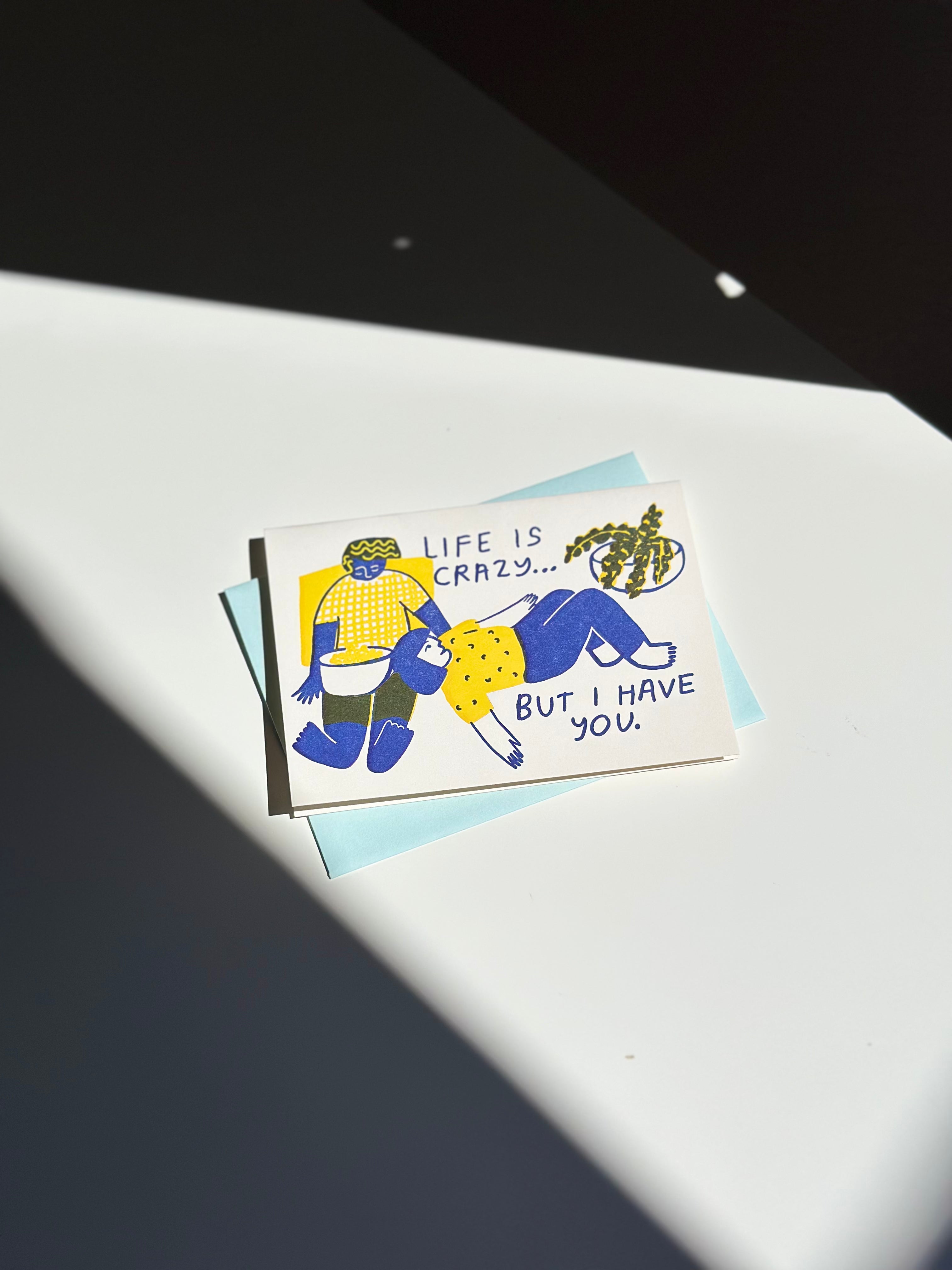 Illustrated greeting card with two people in blue and yellow outfits lounging together, text reads ‘Life is crazy… but I have you,’ paired with light blue envelope.
