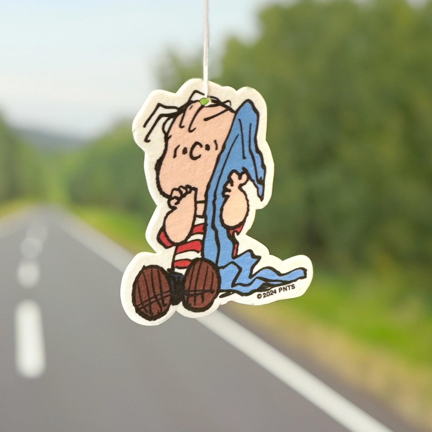 Linus and blanket air freshener hanging in front of a tree-lined road. Cozy Peanuts scent accessory for sentimental and nostalgic car decor.