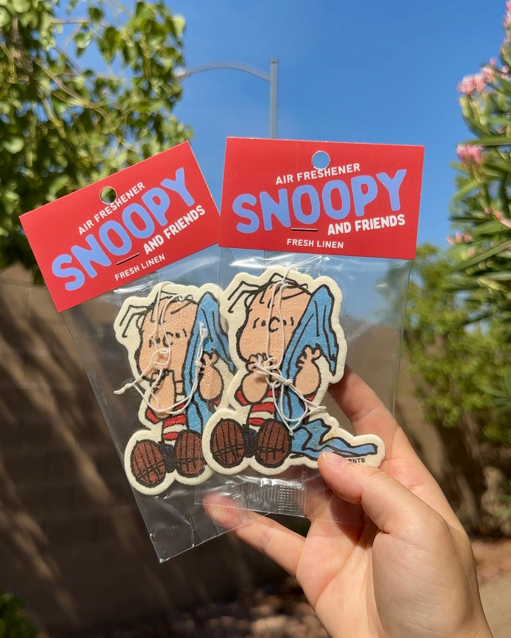 Hand holding two packaged Linus Fresh Linen air fresheners with Linus holding his blue blanket against a sunny outdoor background.