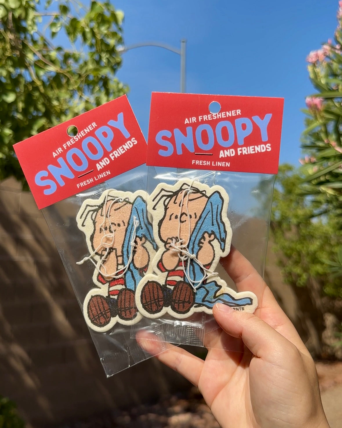 Hand holding two packaged Linus Fresh Linen air fresheners with Linus holding his blue blanket against a sunny outdoor background.