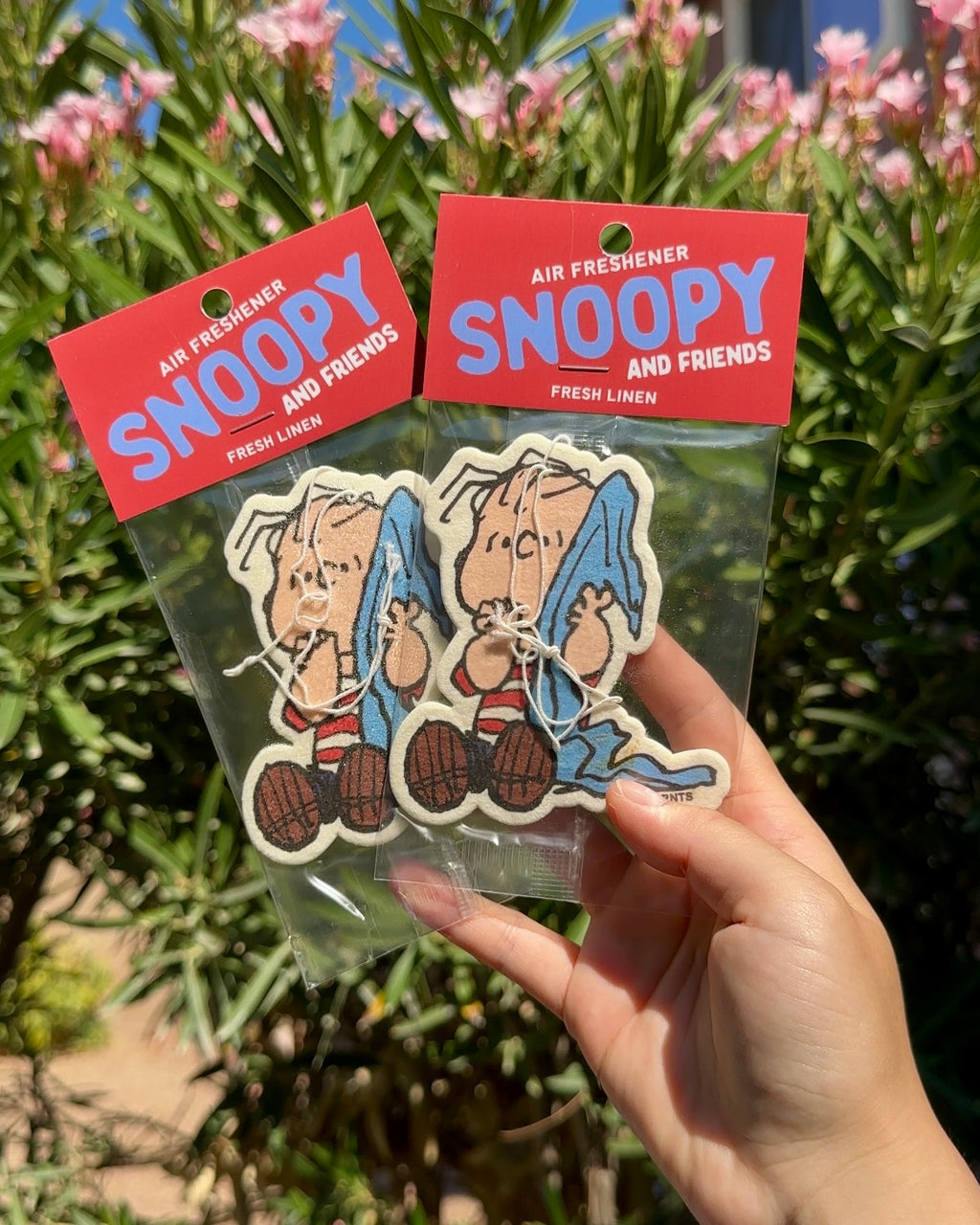 Hand holding two packaged Linus Fresh Linen air fresheners against a green foliage background.