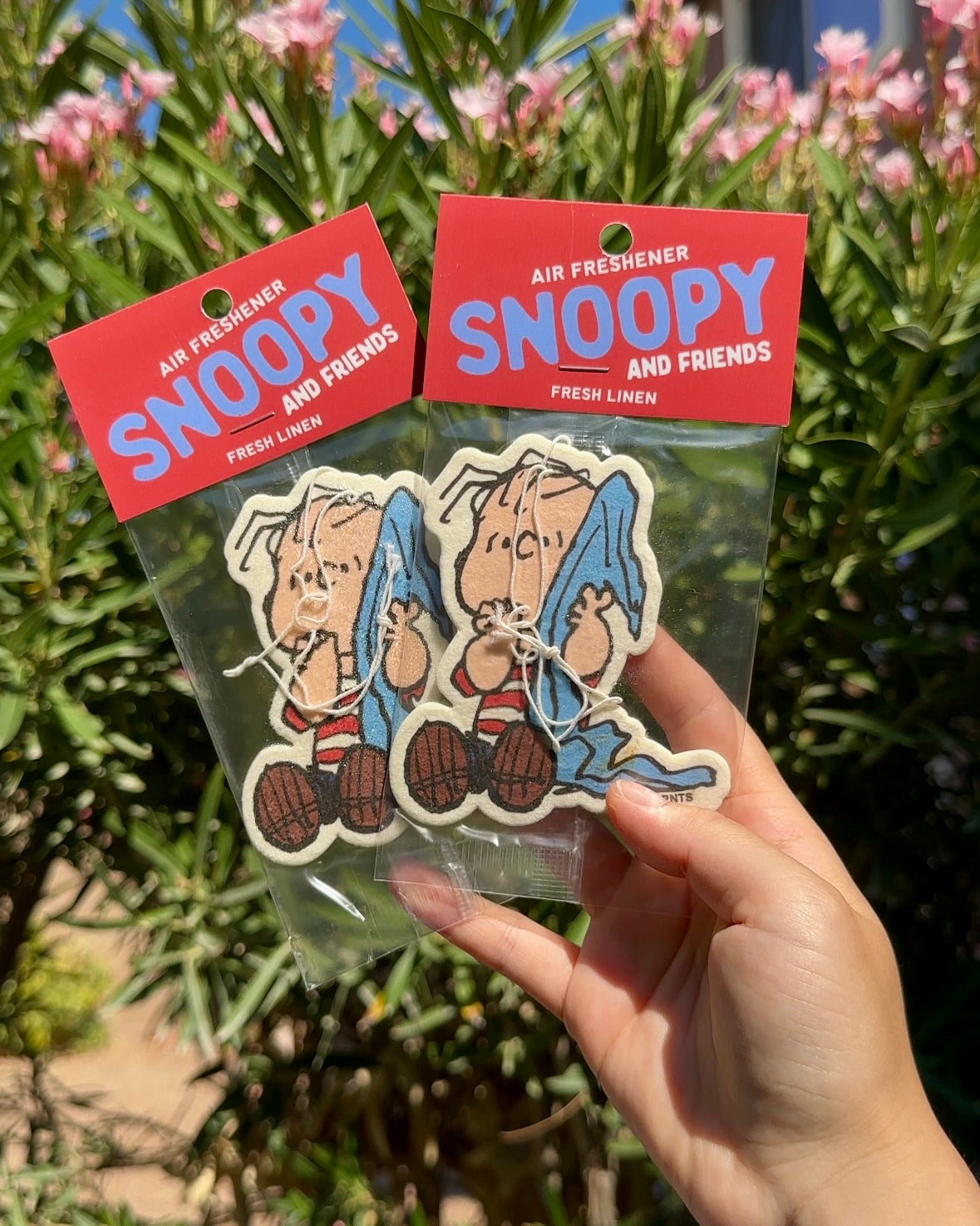 Hand holding two packaged Linus Fresh Linen air fresheners against a green foliage background.