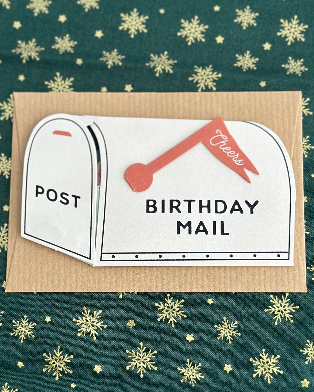 Mailbox birthday card on kraft backing with red “Cheers” flag and the words “BIRTHDAY MAIL,” styled on green fabric with gold snowflakes.