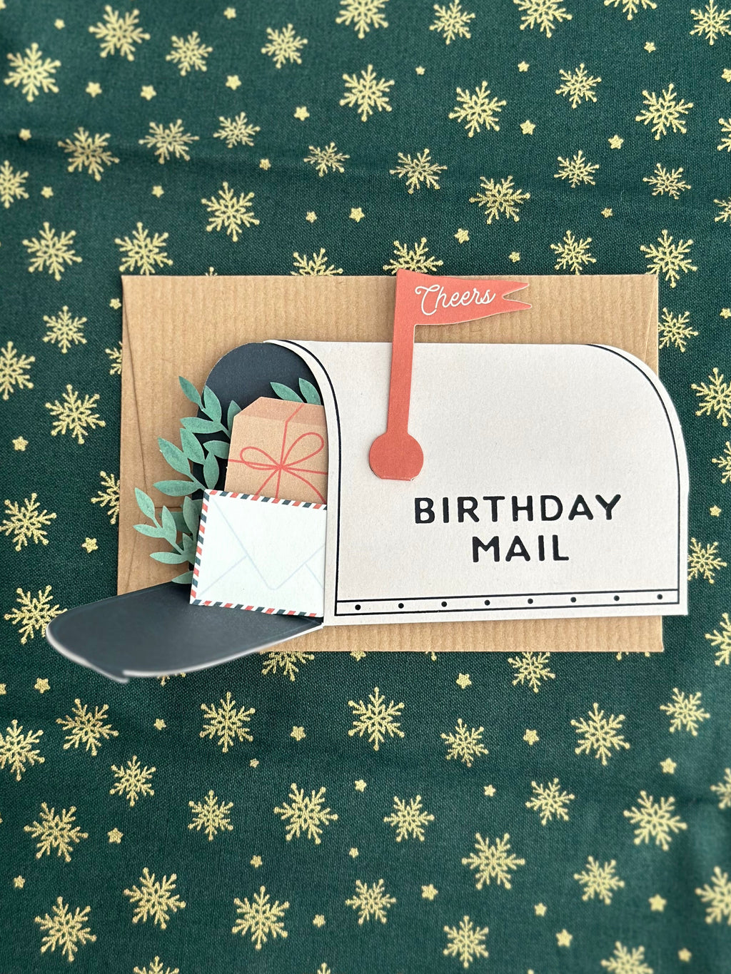 Mailbox birthday card fully opened showing wrapped present, greenery, and small envelope tucked in the mailbox, kraft card base behind.