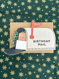 Mailbox birthday card fully opened showing wrapped present, greenery, and small envelope tucked in the mailbox, kraft card base behind.