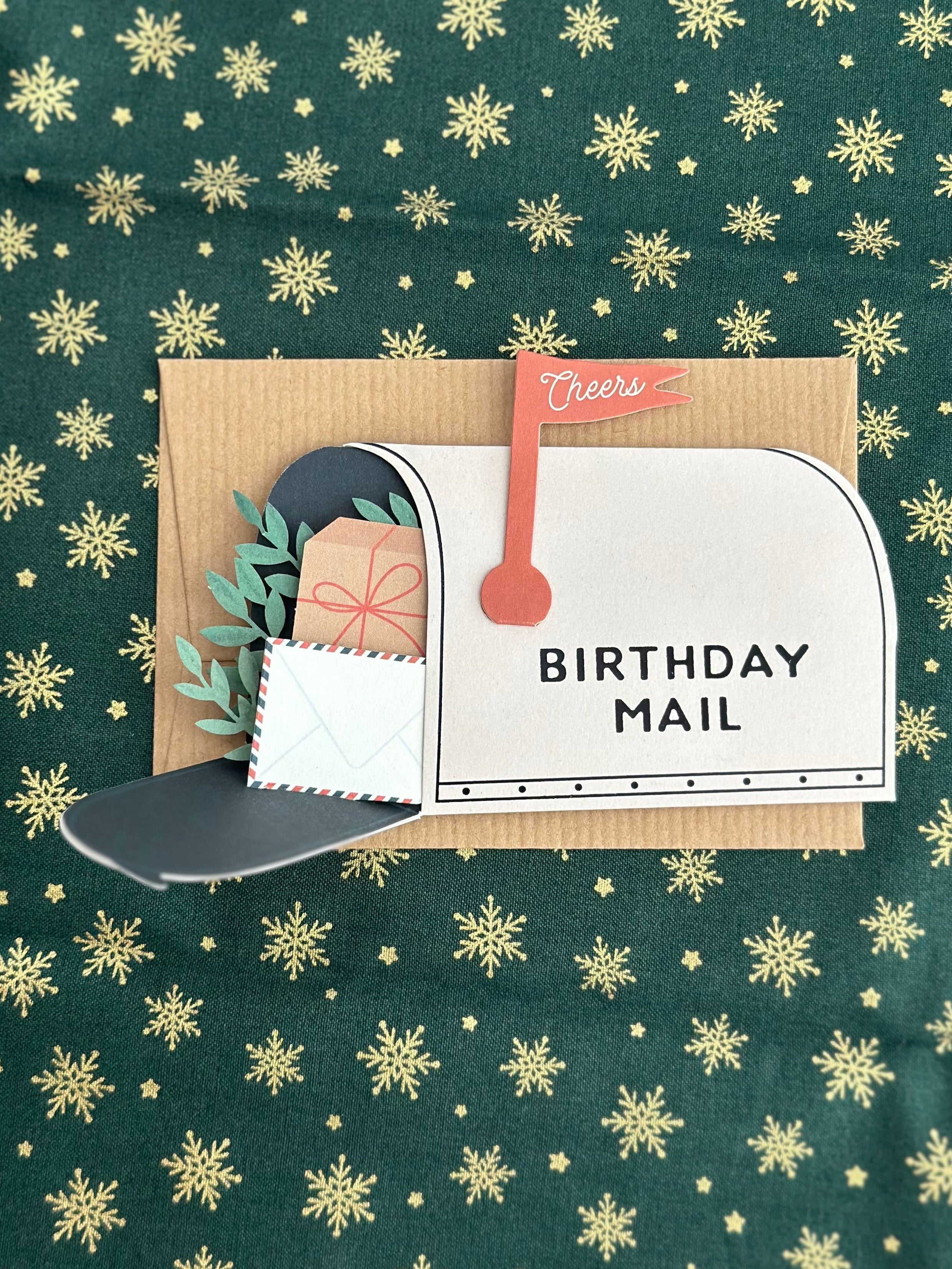 Mailbox birthday card fully opened showing wrapped present, greenery, and small envelope tucked in the mailbox, kraft card base behind.