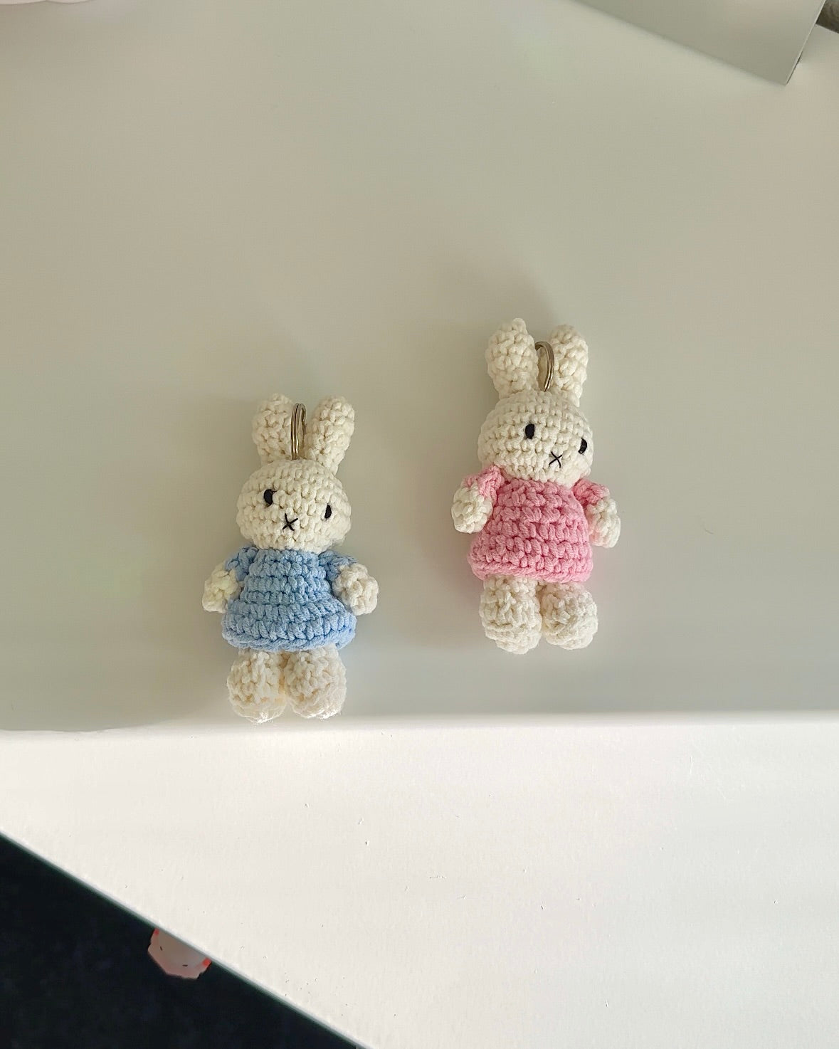 Two Miffy crochet keychains laid flat, one in a baby blue dress and one in a pink dress, handmade cotton bag charms with soft texture.