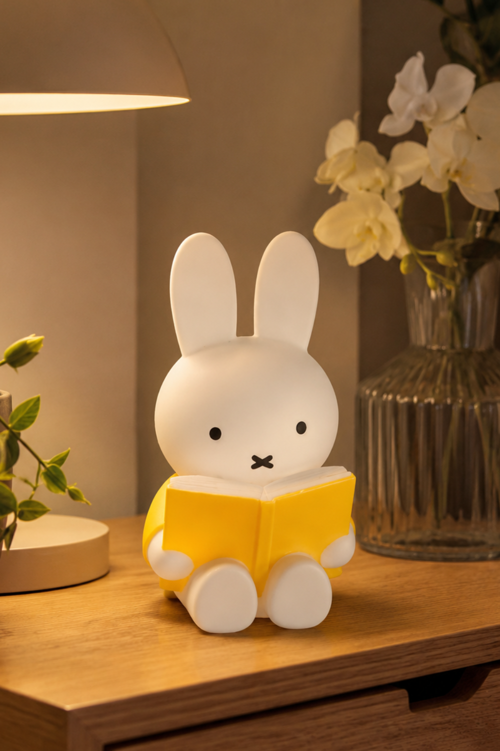 Miffy coin bank with yellow book glowing in warm lamp light on a wooden nightstand, a cozy minimalist bedroom decor piece.
