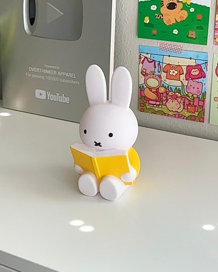 Cute white Miffy figurine reading yellow book, used as coin bank or display