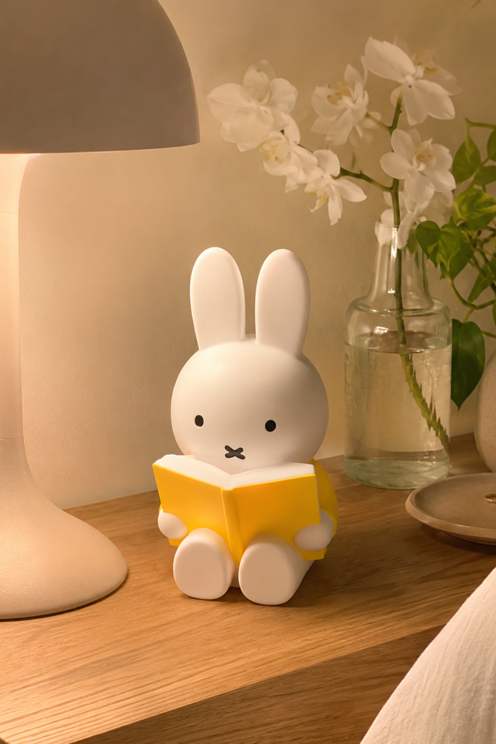 Miffy coin bank with yellow book sitting on a wooden nightstand beside a lamp and white orchids, a cute minimalist bedside decor piece.
