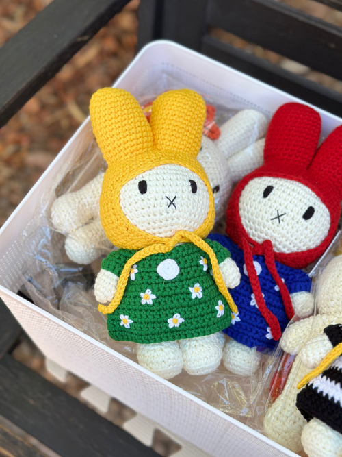 Four handmade crochet Miffy bunny dolls in colorful outfits, including green daisy dress, blue dress with red hood, and black-and-white striped dress with yellow hood, arranged in a white basket.