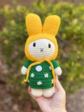 Close-up of handmade crochet Miffy bunny in yellow hood and green daisy dress, held outdoors against a blurred background.