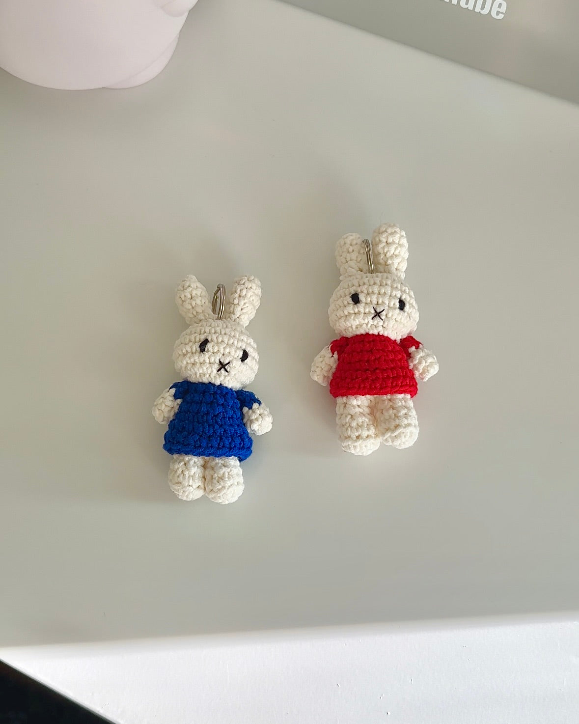 Two Miffy crochet keychains in red and blue dresses laid flat on a light surface, handmade cotton Miffy bag charms with metal keyrings.