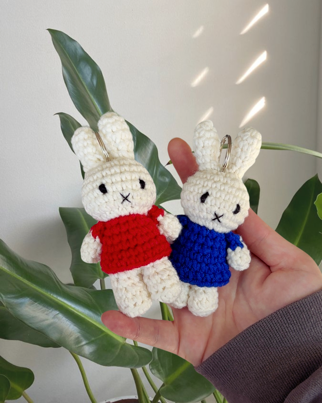 Hand-crocheted Miffy keychain in a red dress, made from cotton yarn and held in hand with green plant background, soft and cozy Miffy bag charm.