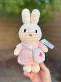 Handmade Miffy crochet plush in a pastel pink dress with a removable blue bag, held outdoors against a blurred background.