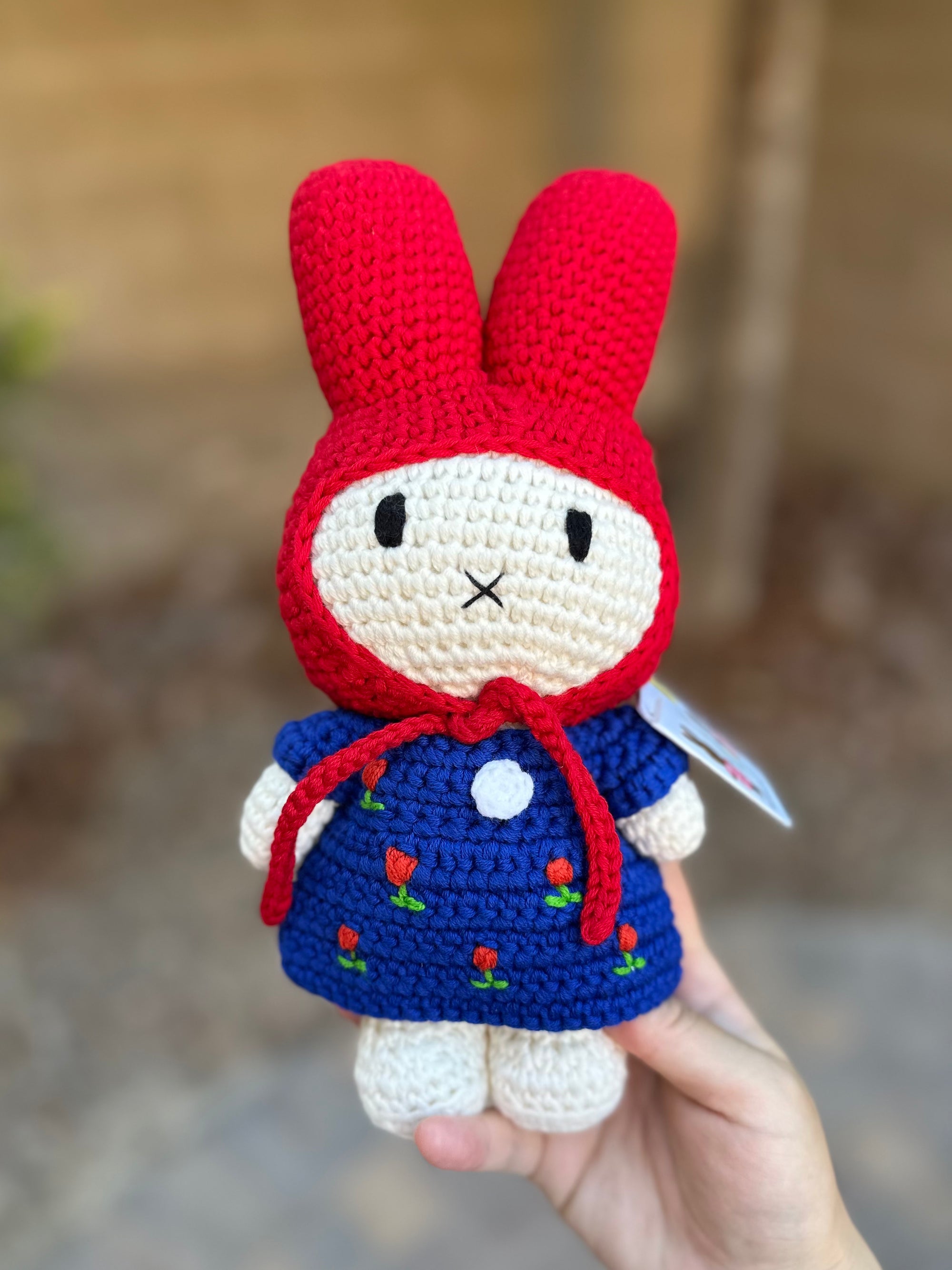 Handmade Miffy crochet doll in a red bonnet and blue tulip dress, held outdoors.