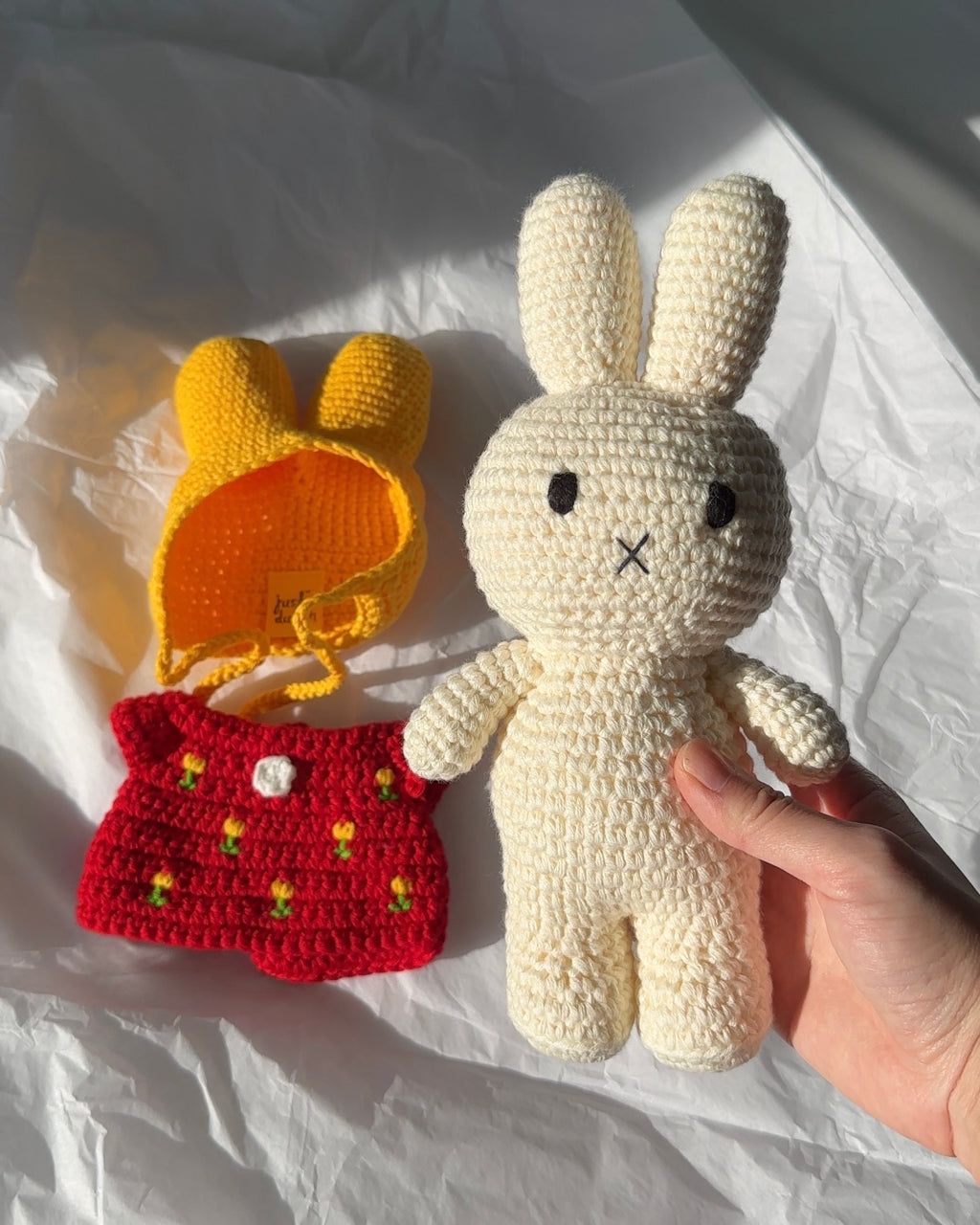 Hand-crocheted Miffy plush by Just Dutch with red tulip dress and yellow bonnet, made from cotton yarn, showing removable outfit pieces on a light background.
