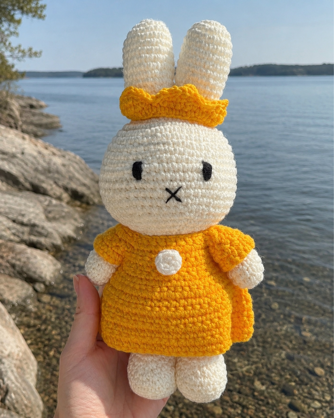 Miffy crochet bunny plush in her Yellow Dress & Crown, held on a rocky lake shore with calm water in the background.