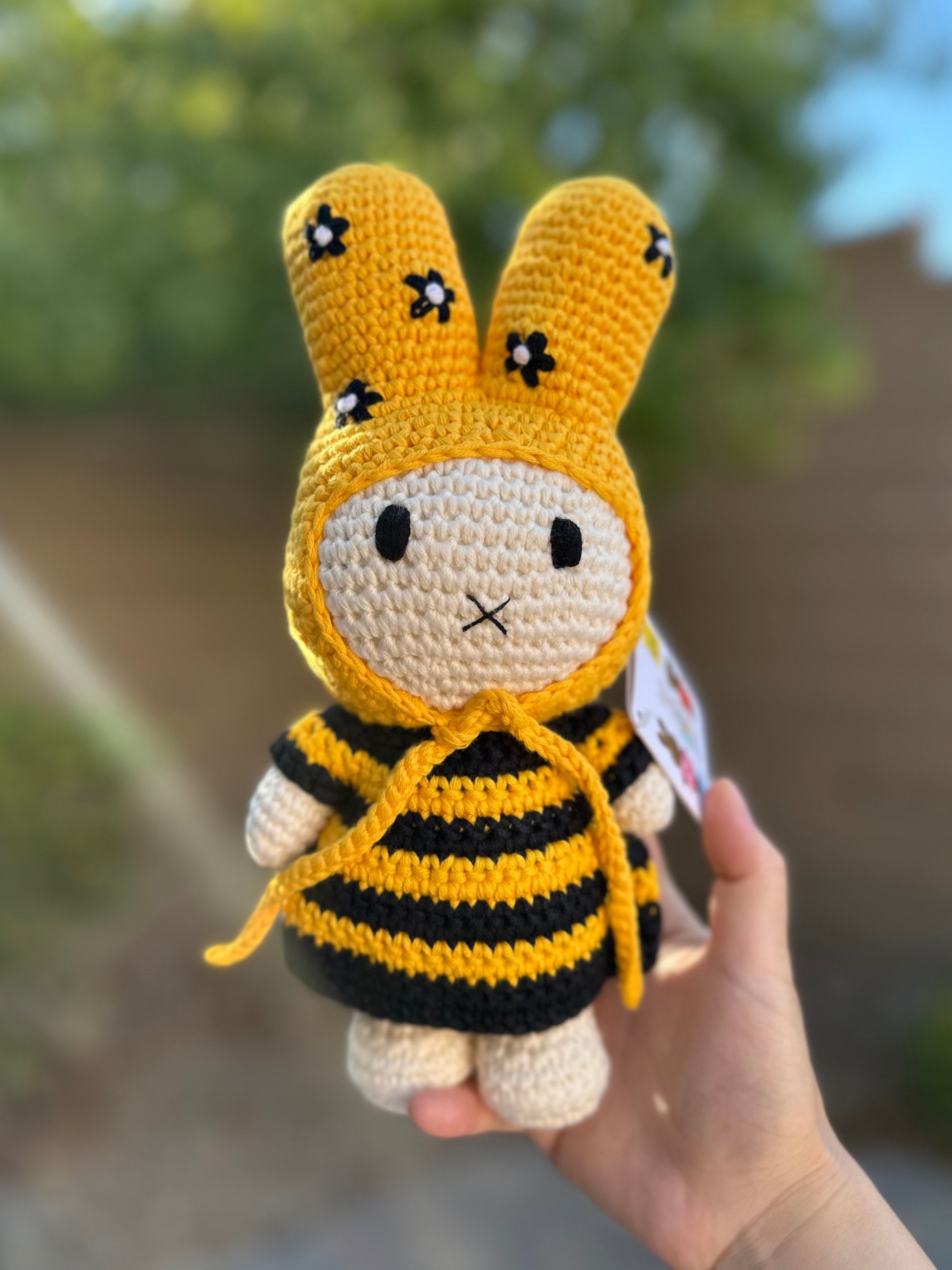 Handmade Miffy crochet plush in a black-and-yellow bee dress with a yellow floral bonnet, held outdoors against a blurred background.