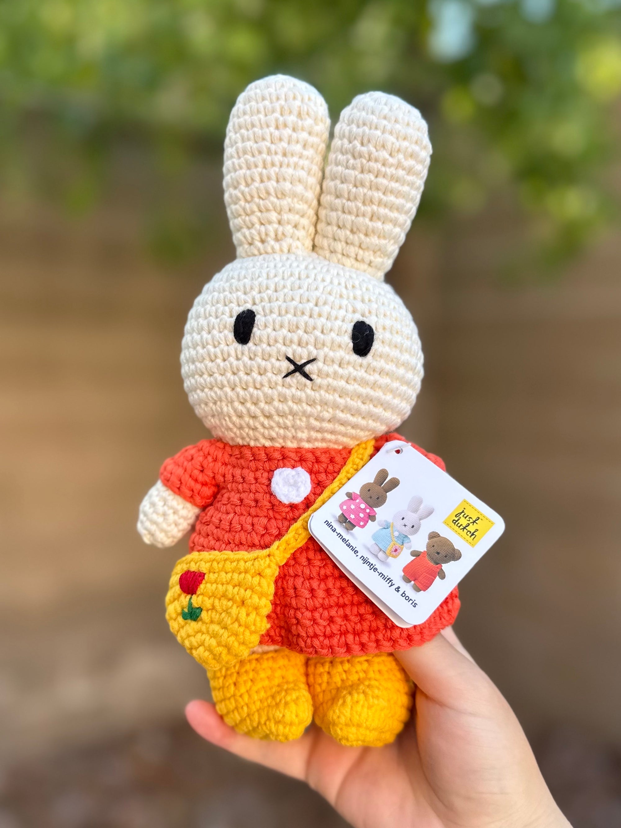 Handmade Miffy crochet plush in a red dress with yellow shoes and a tulip-embroidered crossbody bag, held outdoors against a blurred background.